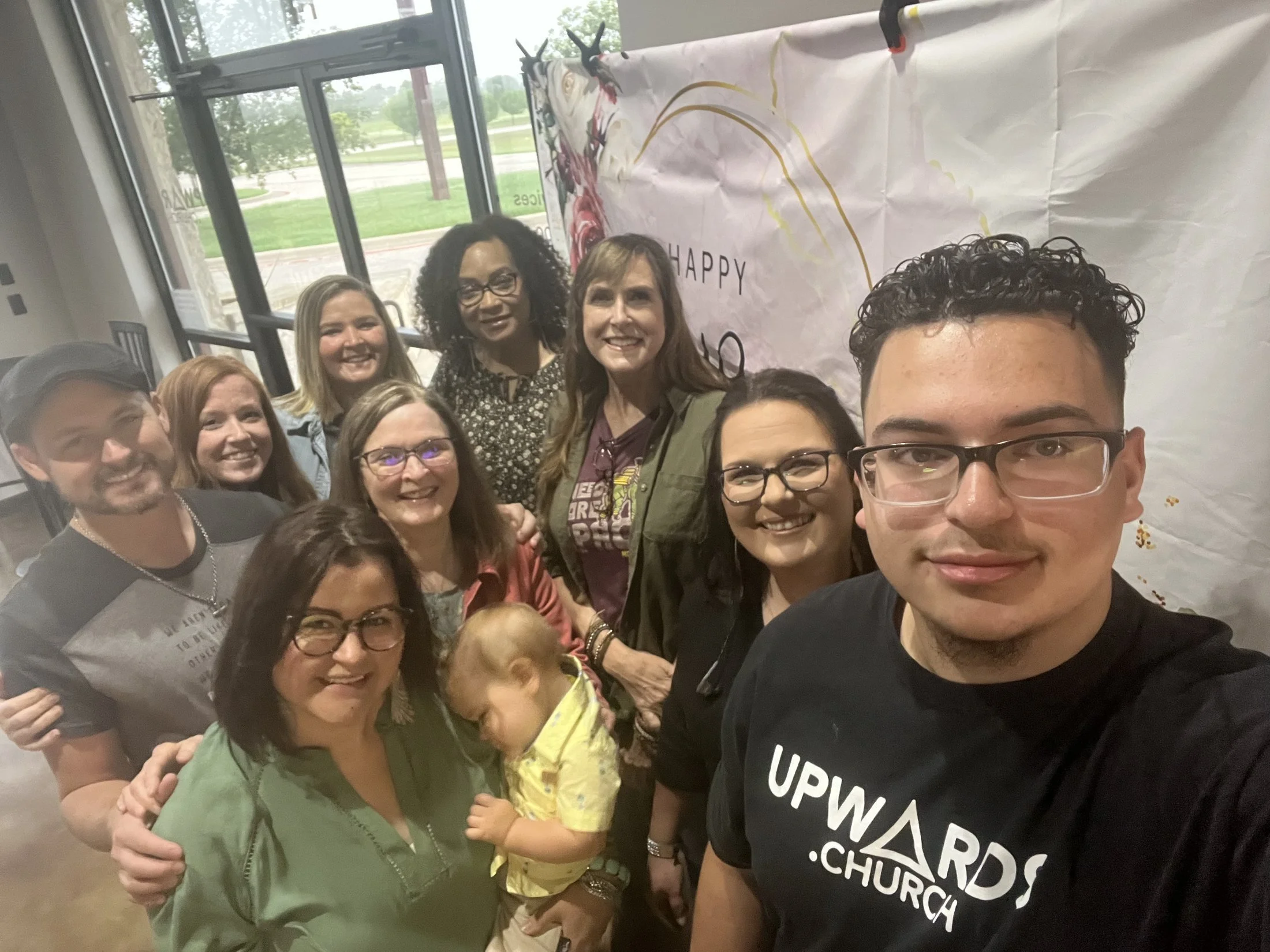 A group of people, including women and children, standing together indoors for a group photo. There are large windows and a banner with the word 'HAPPY' in the background.