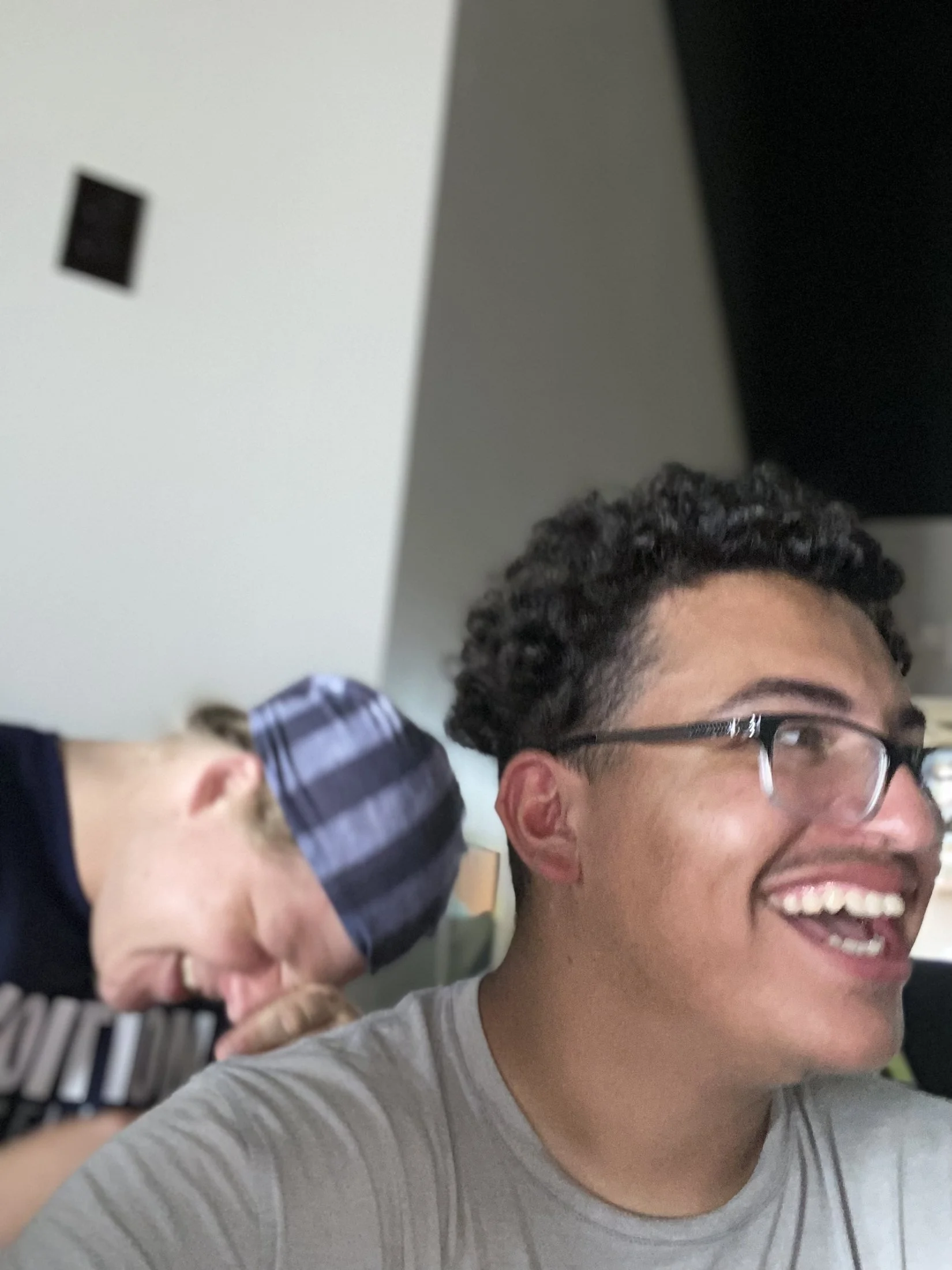 Close-up of a young man with glasses smiling, wearing a gray shirt, and holding a striped object, with part of another person's head and hand in the background.