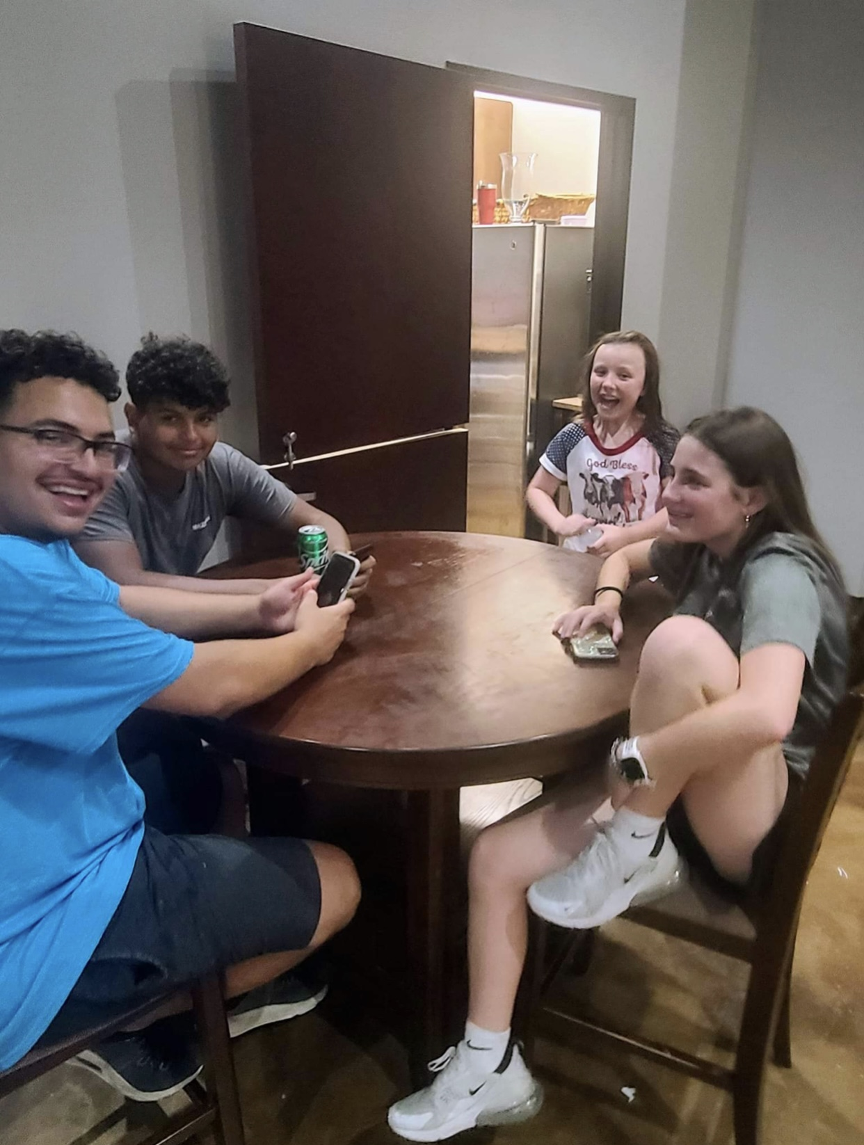 Five people sitting around a wooden table laughing and chatting, two girls and three boys, one girl with a phone on her lap, one boy with a soda can, and the others holding phones.