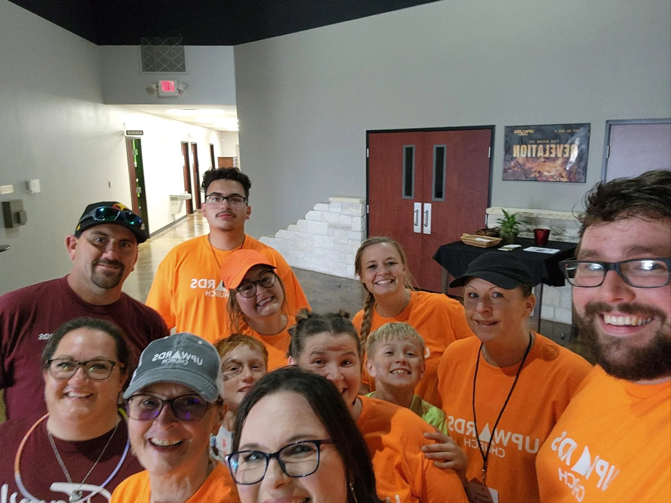 Group of people, including men, women, and children, smiling and posing together indoors. Some individuals are wearing orange shirts, and one person has a bicycle helmet. The background shows a hallway with a red door, posters on the wall, and tables