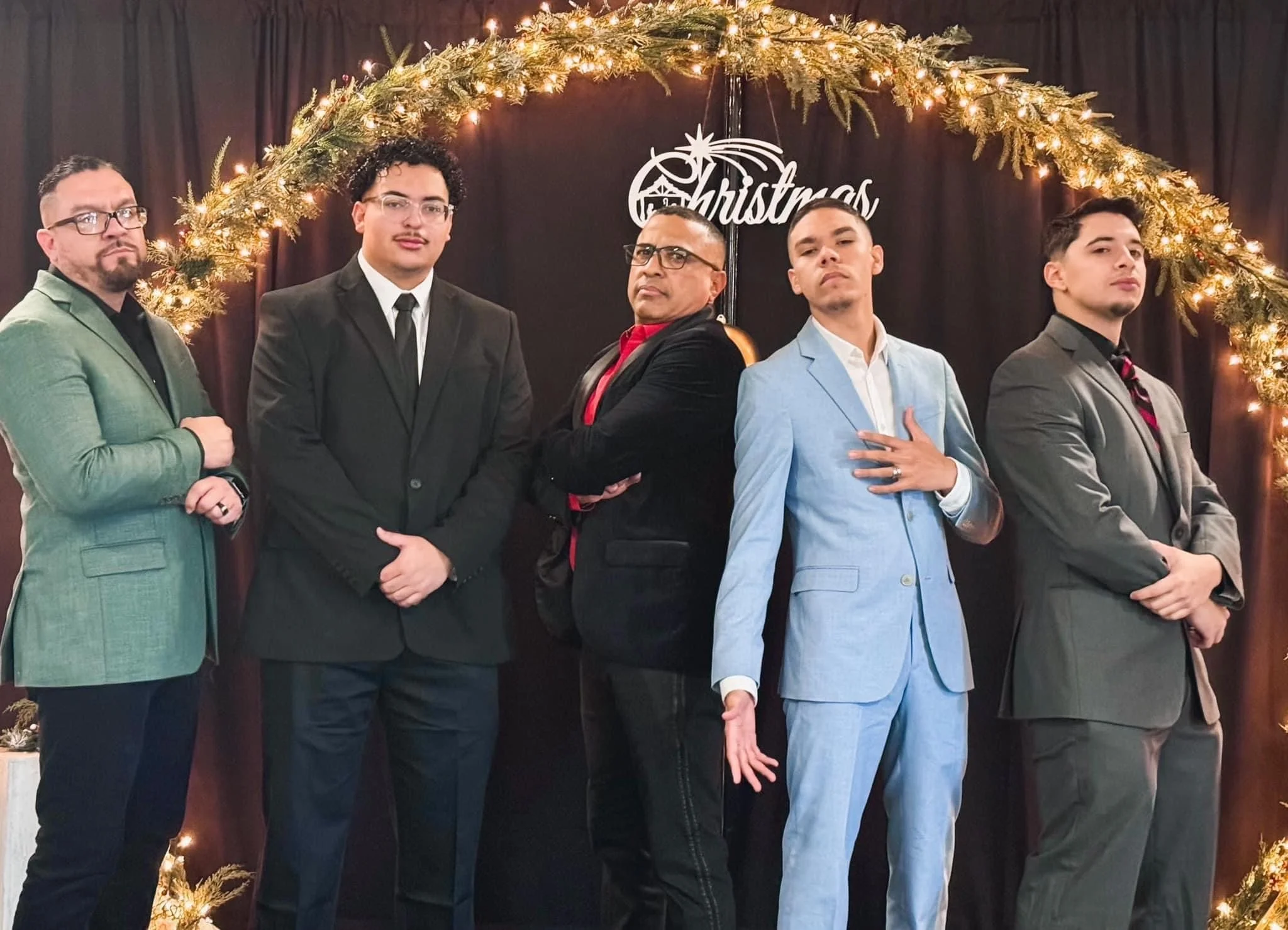 Five men stand in a line in front of a dark backdrop with a Christmas decoration arch made of pine branches and lights, and a sign that says 'Christmas'. They are dressed in formal and semi-formal attire, with serious expressions.