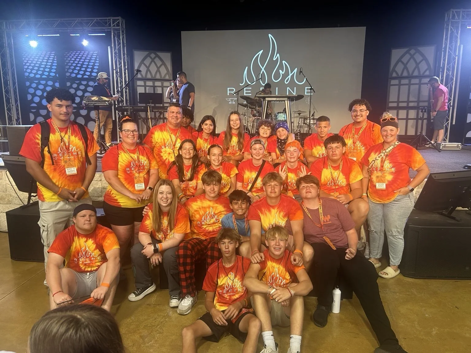 Group of children and teenagers in matching tie-dye shirts with flames, posing for a photo in front of a stage at a conference or event called REDEFINE.