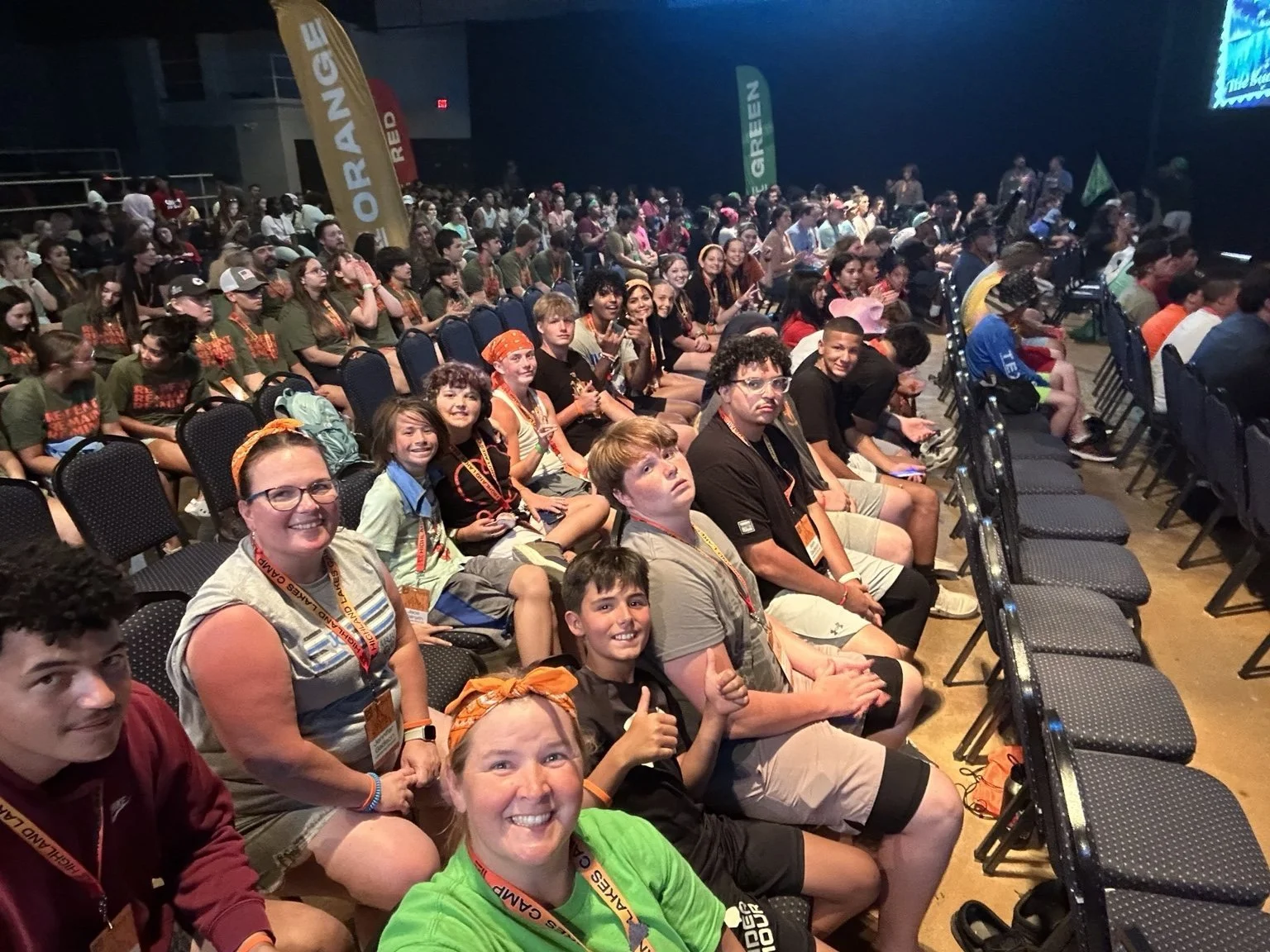 A large group of children and adults sitting in an auditorium, attending an event or conference, with some wearing orange bandanas and lanyards.