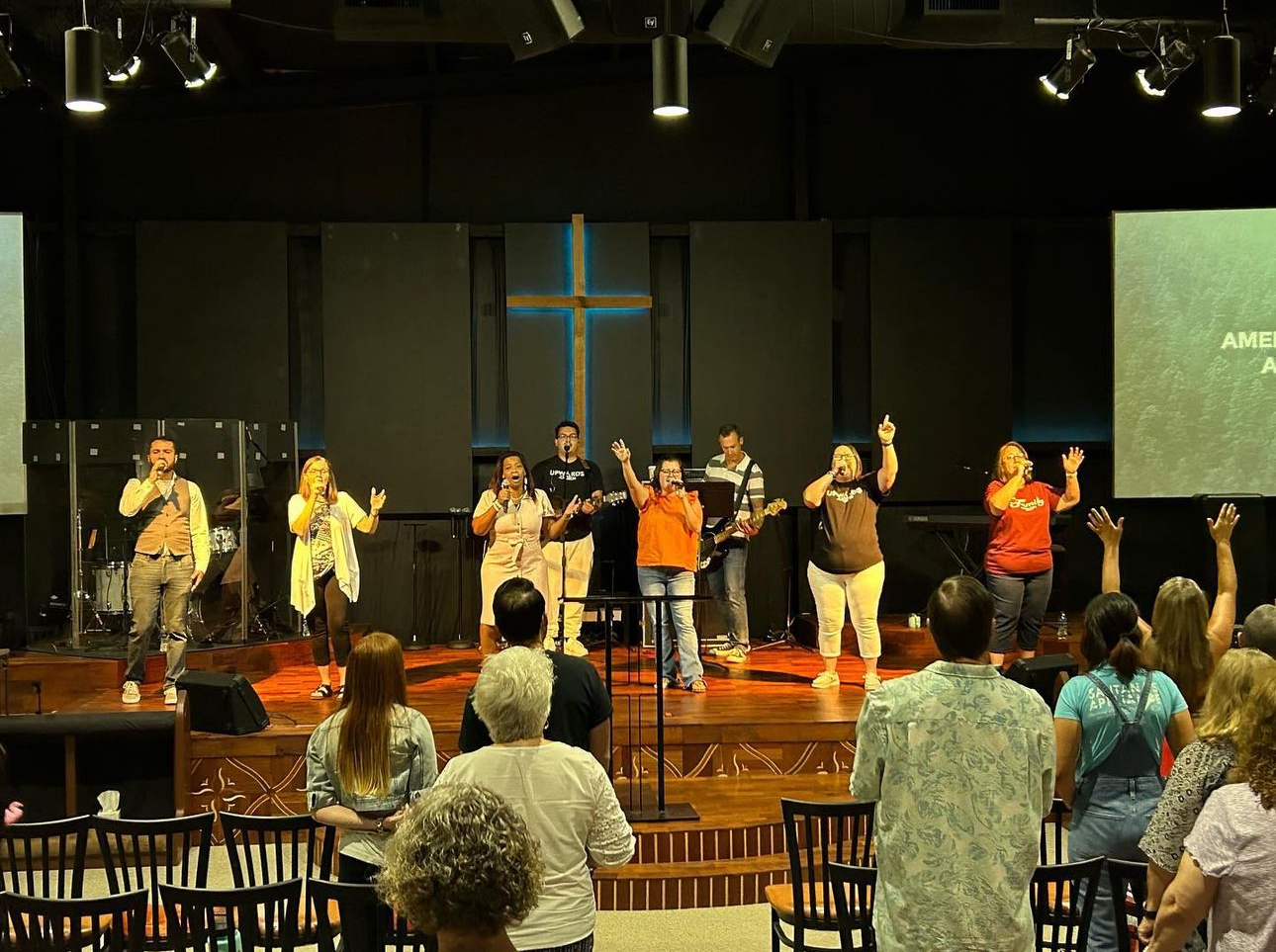 A group of seven singers and musicians performing on a church stage with a large wooden cross in the background, and congregation members with raised hands in front.