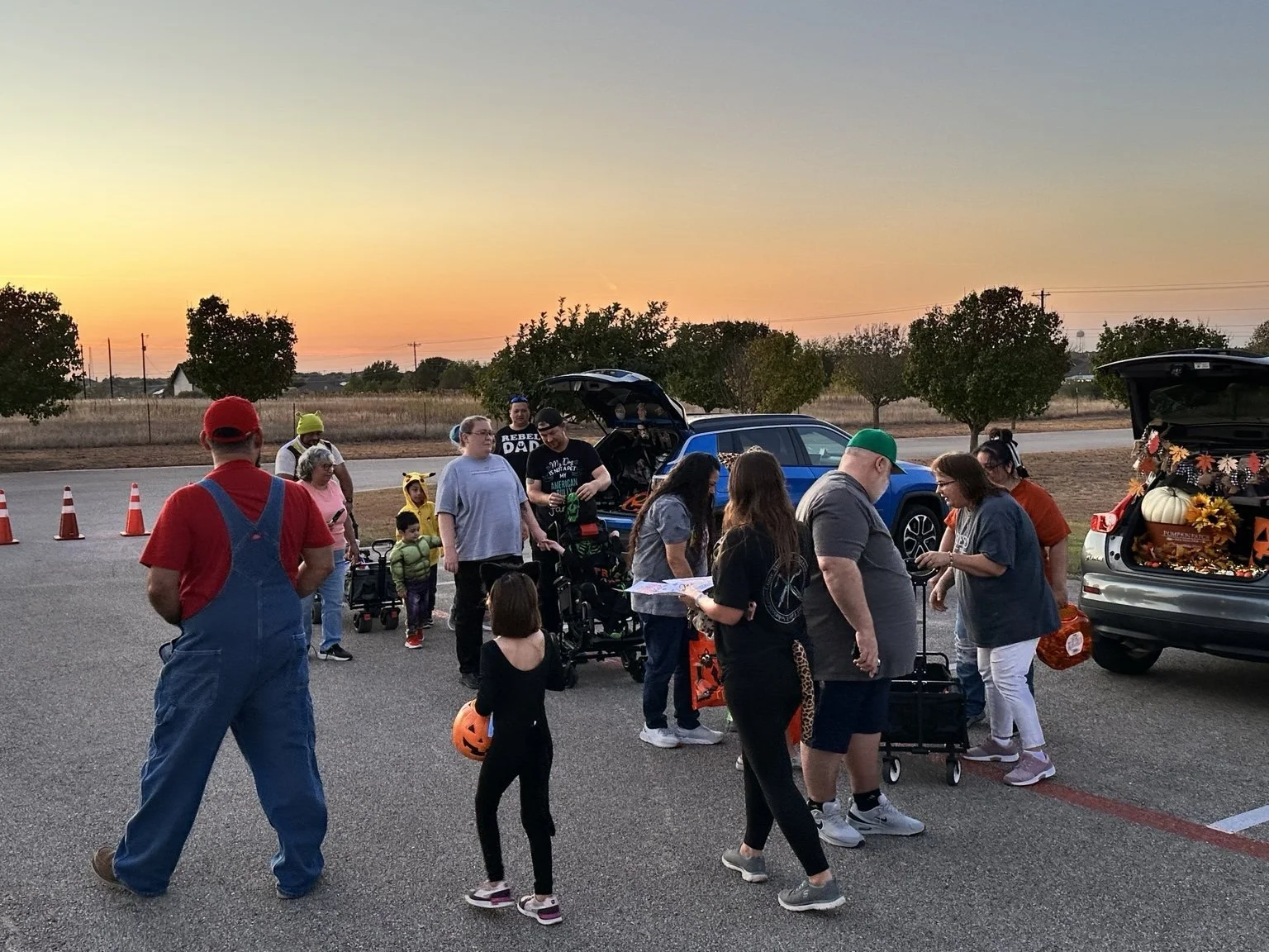 People gather in a parking lot during sunset, with some children holding Halloween buckets and dressed in costumes. Cars have open trunks decorated for Halloween, and orange traffic cones are set up in the background.