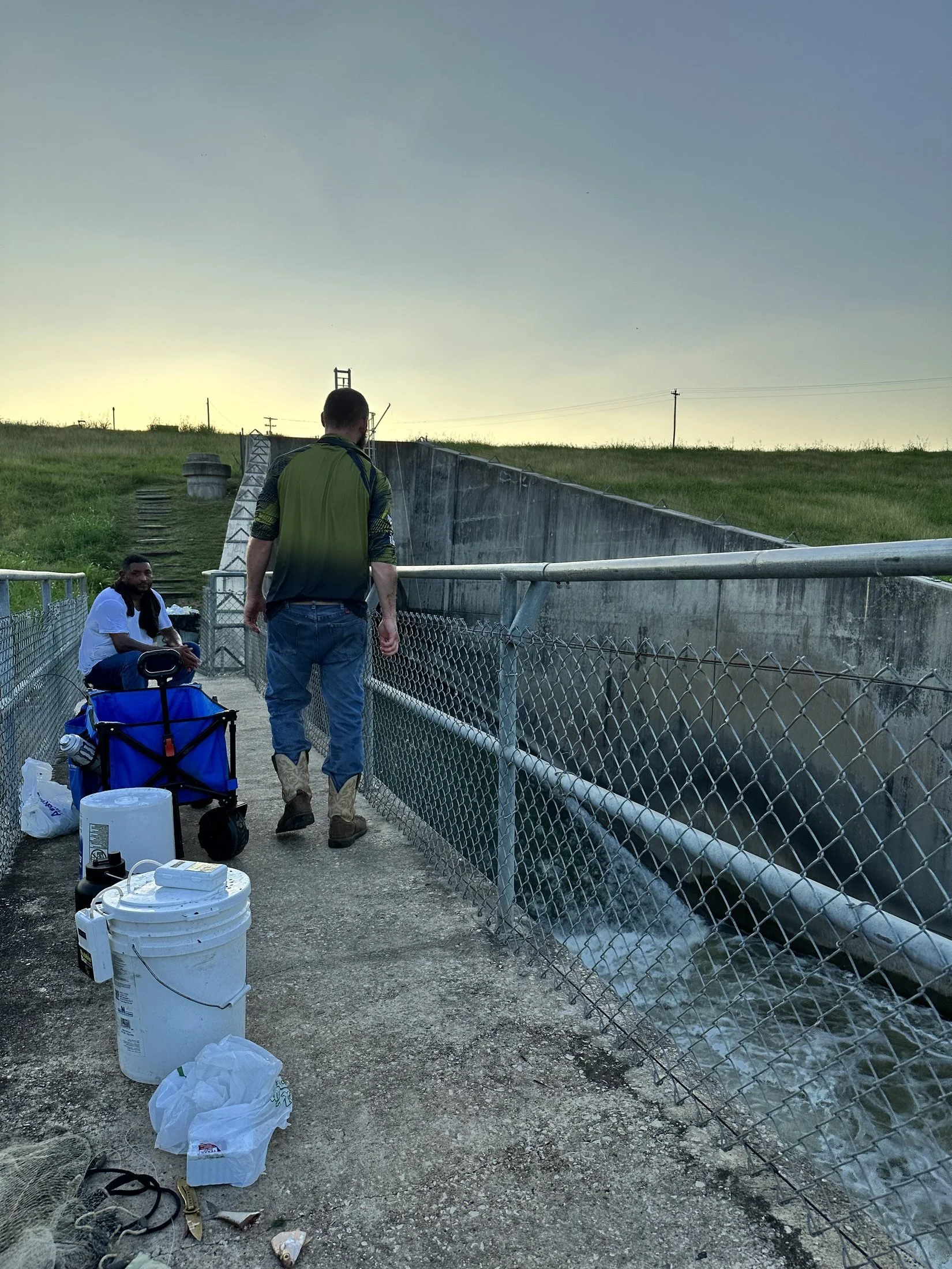 Two men by a chain-link fence near a body of water with a grassy hill in the background. One man is sitting, wearing a white shirt, and the other man is walking away, wearing a green and black shirt. Various items including buckets, bags, and tools a