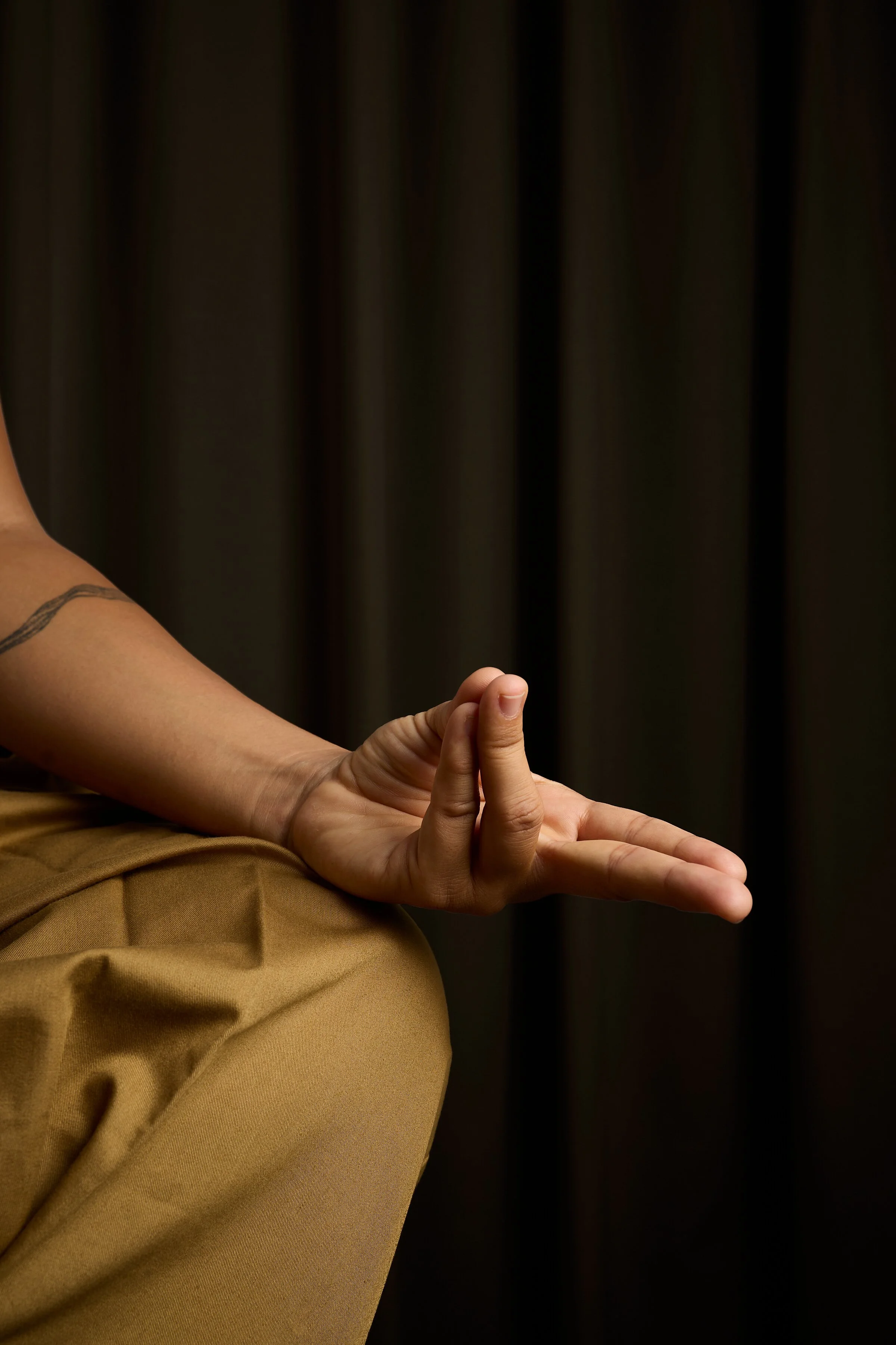 A person performing a meditative yoga pose with their hand resting on their knee, fingers in a mudra, against a dark curtain background.
