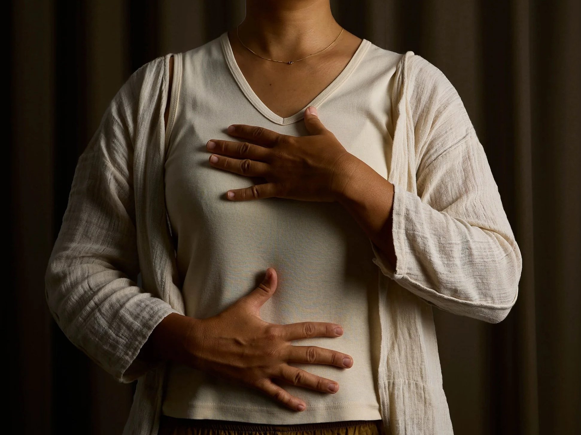 A person wearing a beige sleeveless top and a light-colored cardigan placing one hand on their chest and the other on their stomach, suggesting they may be experiencing discomfort or taking a deep breath.