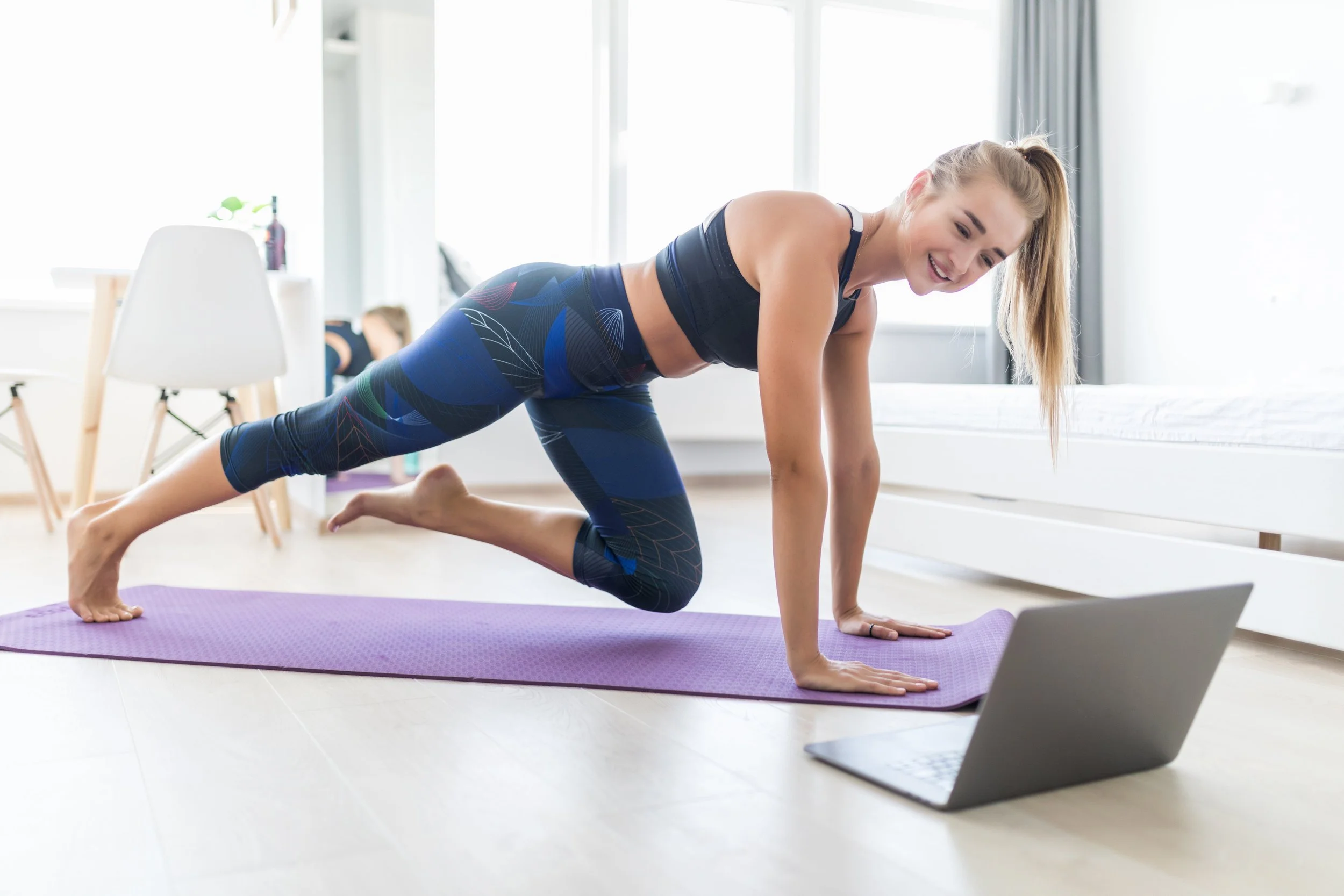 A young woman performing a plank yoga pose on a purple mat in a bright, modern room with large windows, while watching a laptop placed on the floor.