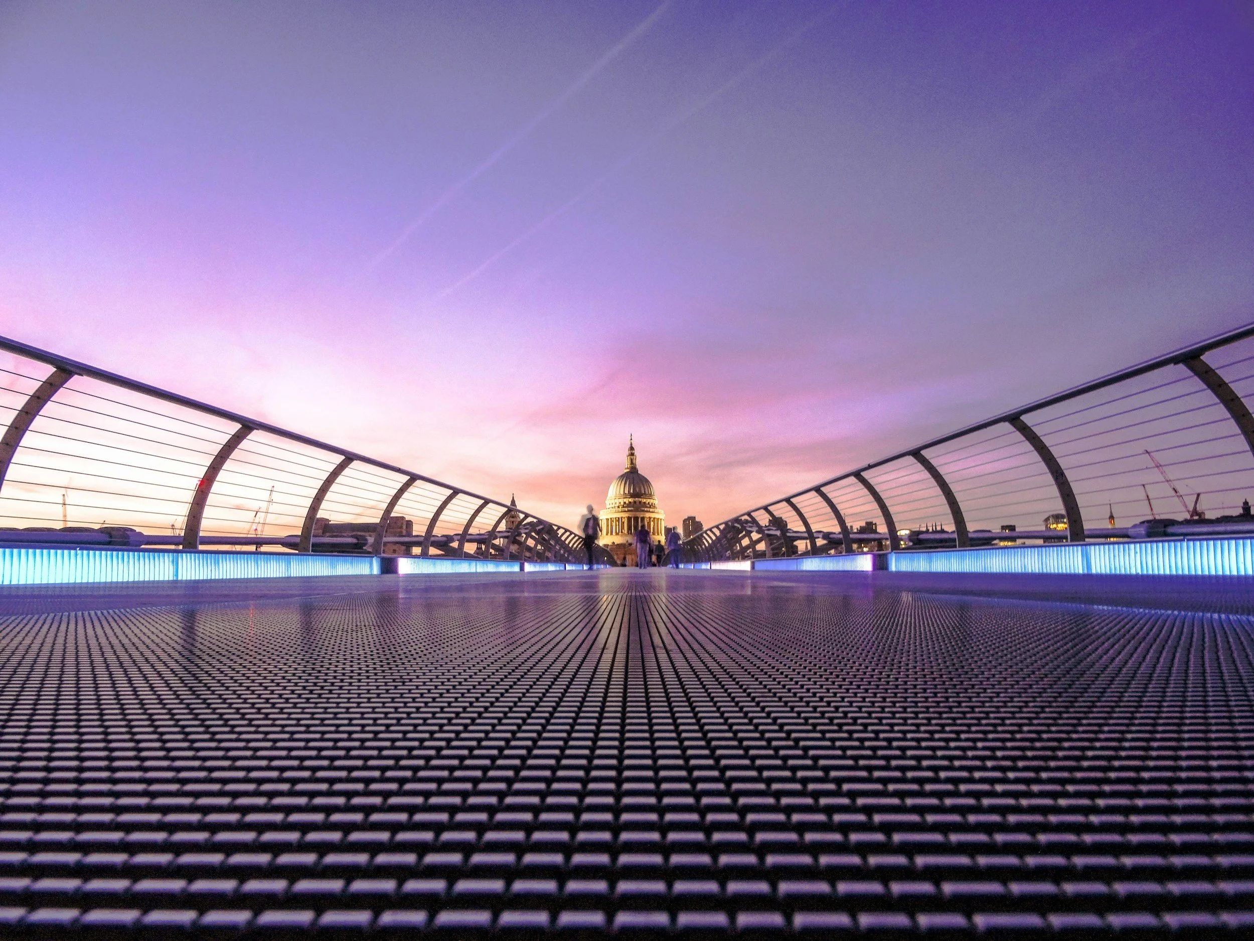 View of St. Paul's Cathedral in London seen from Millennium Bridge during sunset, with purple and pink sky and a modern pedestrian bridge in the foreground.