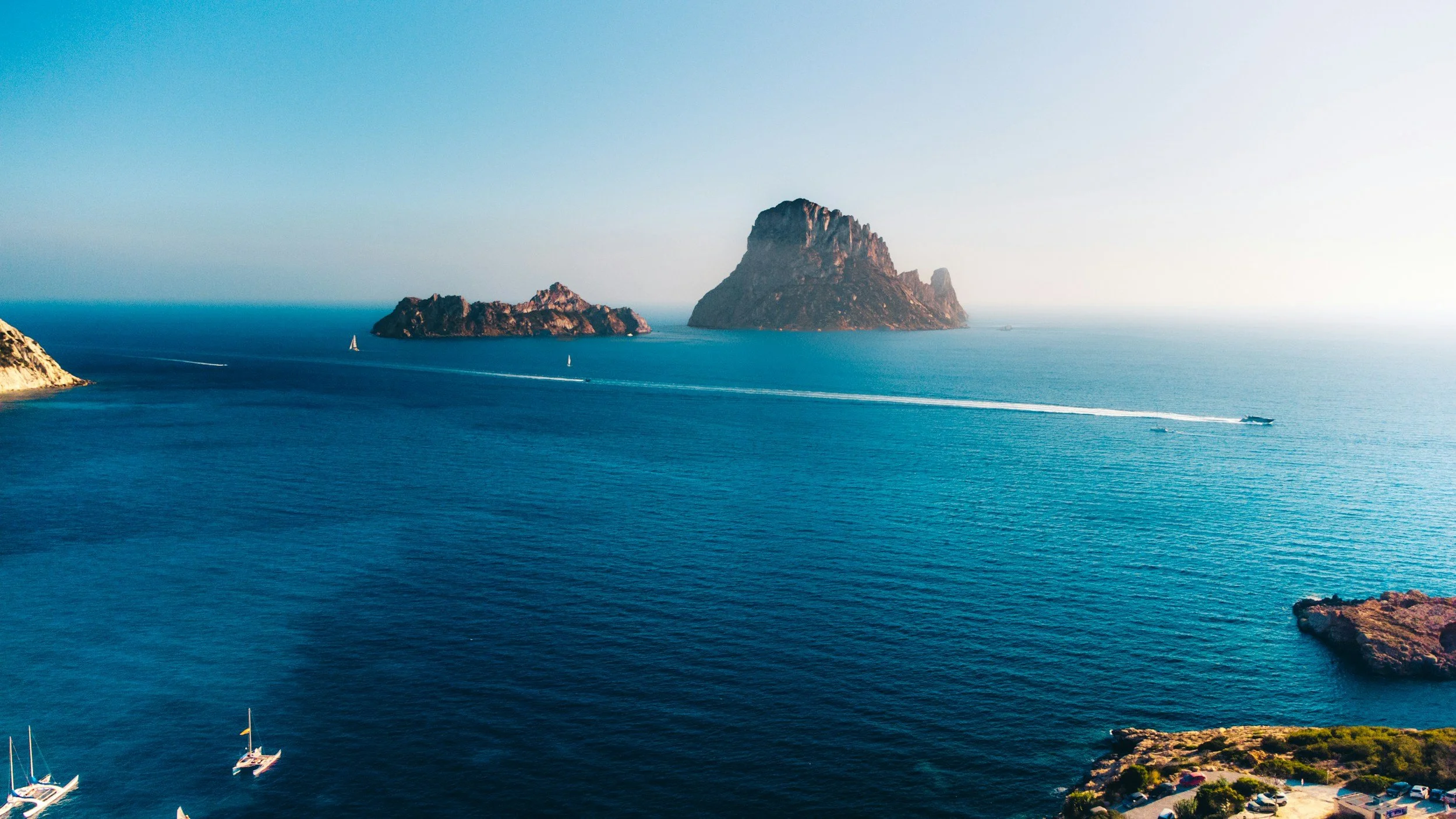 A scenic view of the ocean with several rocks and small islands, sailboats, and a boat leaving a wake in the water, under a clear sky.