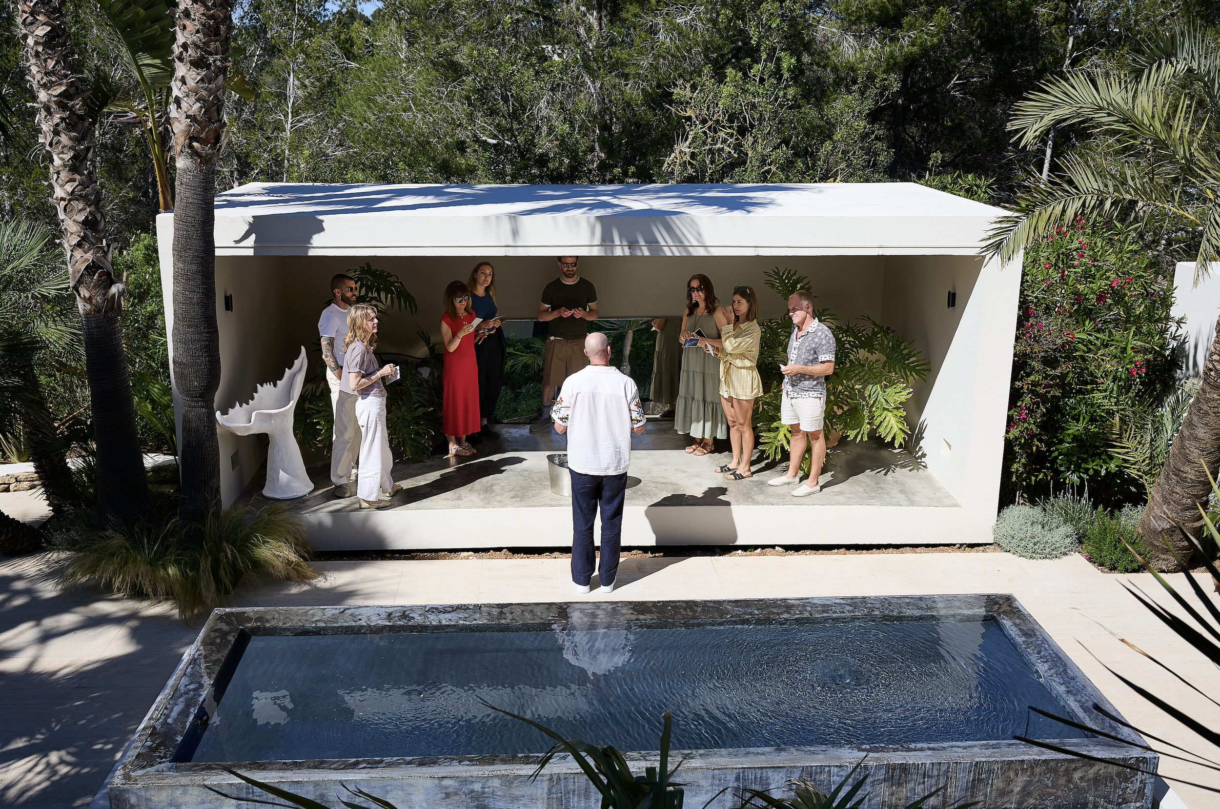 A group of people gathered in an outdoor, modern architectural structure with a white roof, surrounded by lush green tropical plants and trees. The scene appears to be a social or meeting event under a pavilion by a small pool.