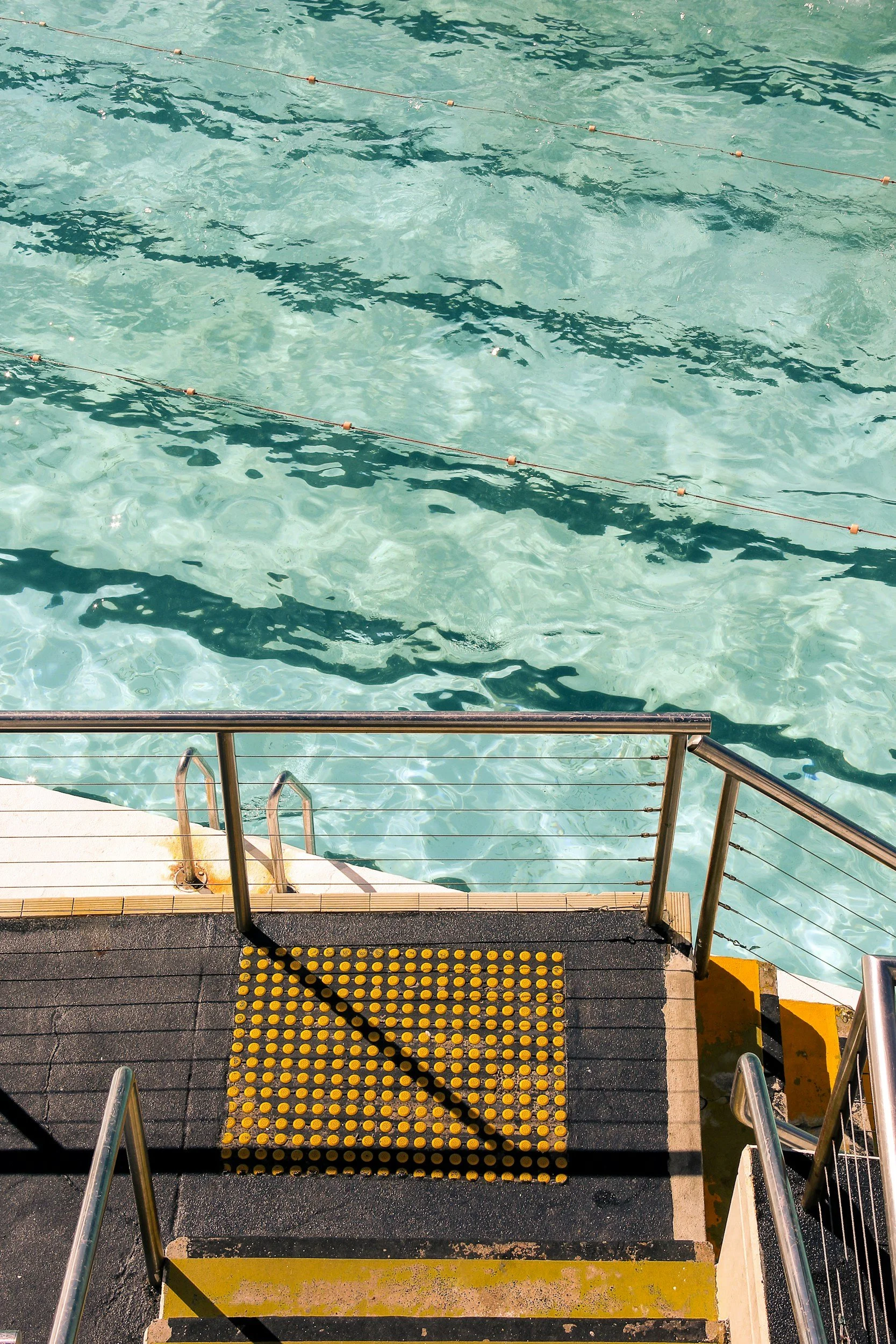 A swimming pool with clear blue water, metal handrails and a staircase leading into the water, surrounded by a black and yellow textured mat and yellow caution lines.