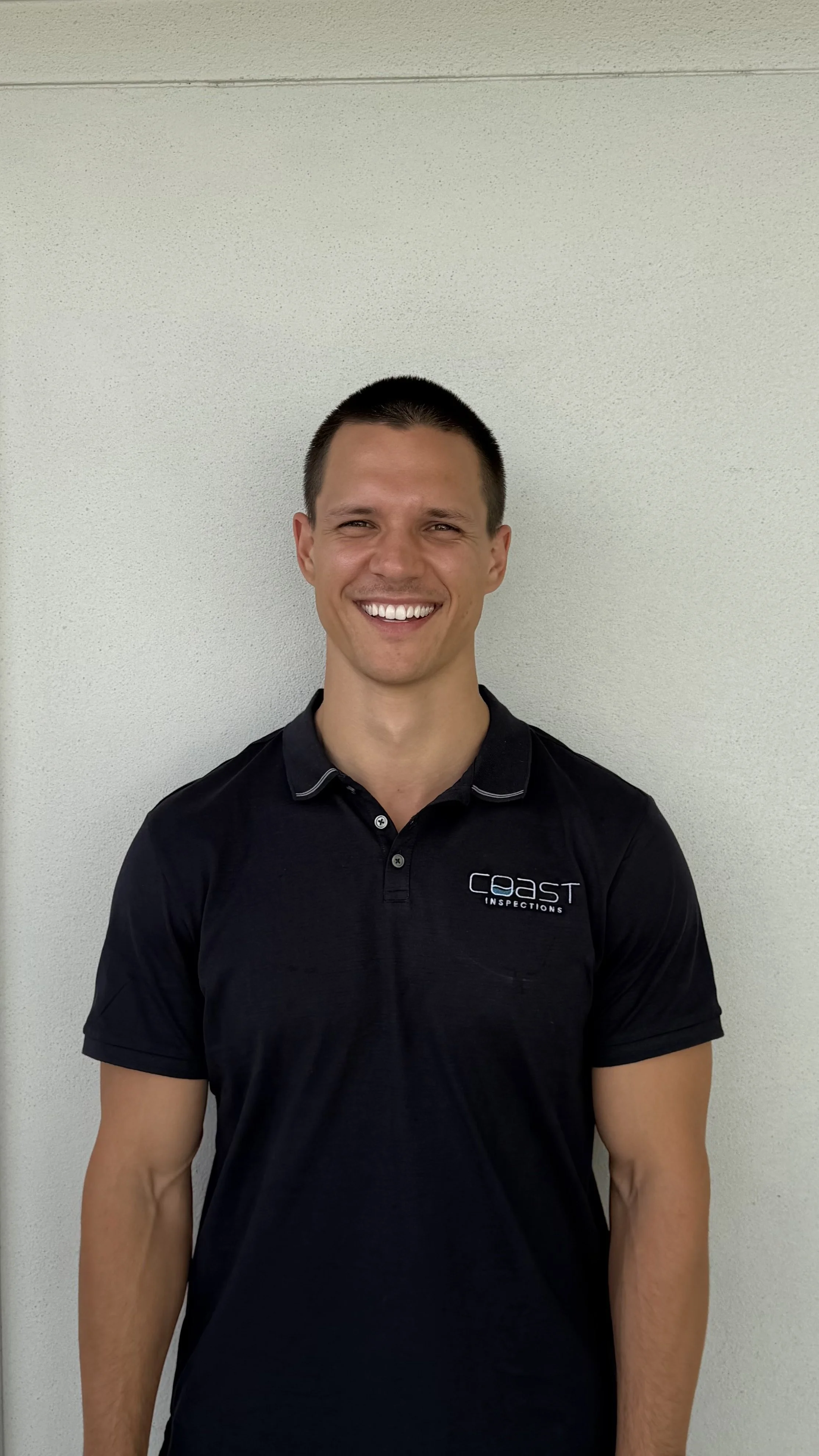 A smiling man with short dark hair in a black polo shirt with a logo that says 'coast inspections' on a plain light-colored wall.