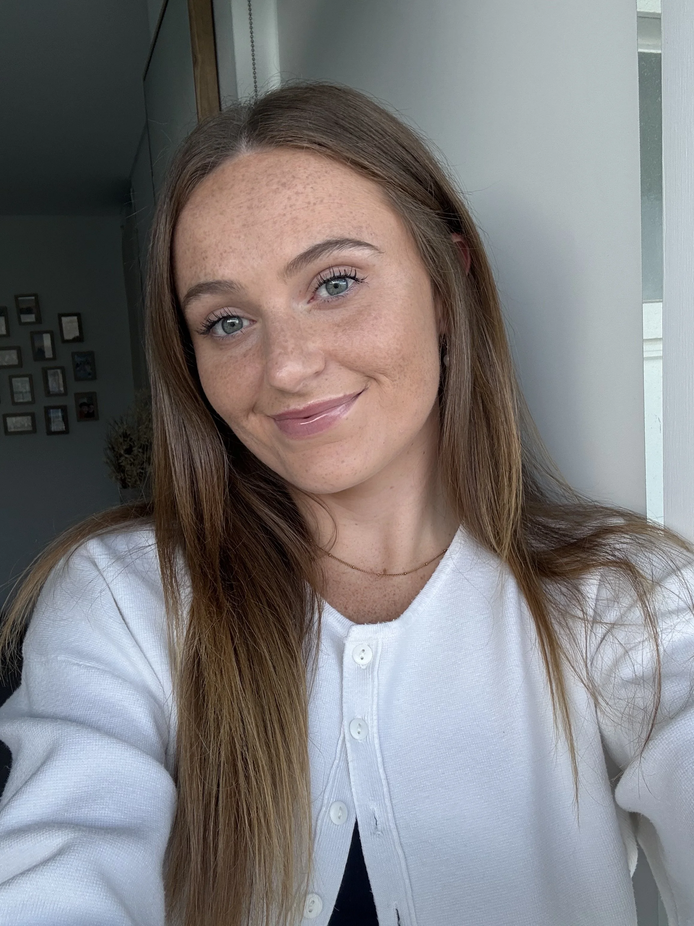 Close-up of a young woman with long, light brown hair, freckles, blue eyes, and a friendly smile, wearing a white button-up shirt and a thin necklace, standing near a window with natural light.