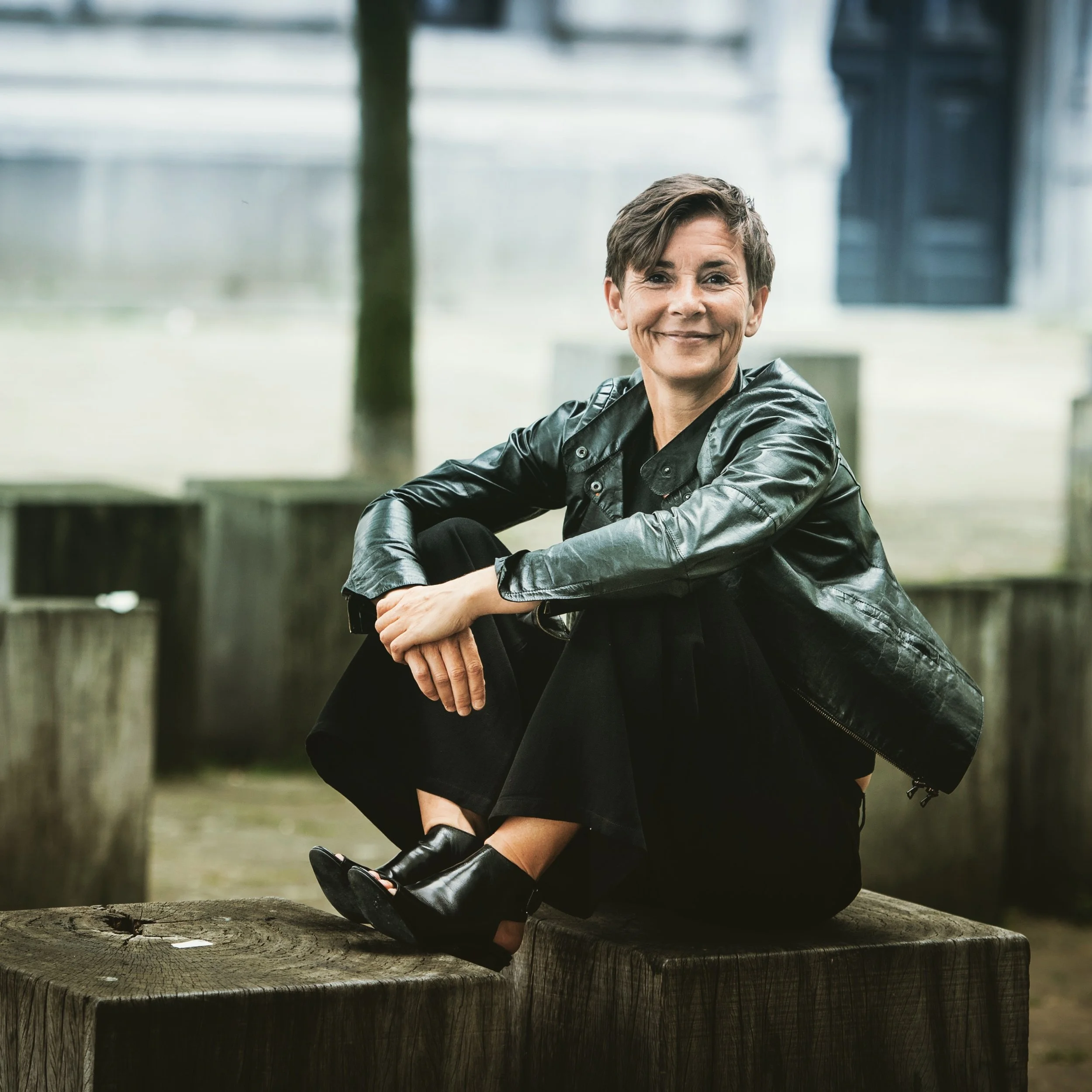 Inge Geerdens, zittend op een houten blok, draagt een zwarte leren jas en laarzen, glimlachend naar de camera.