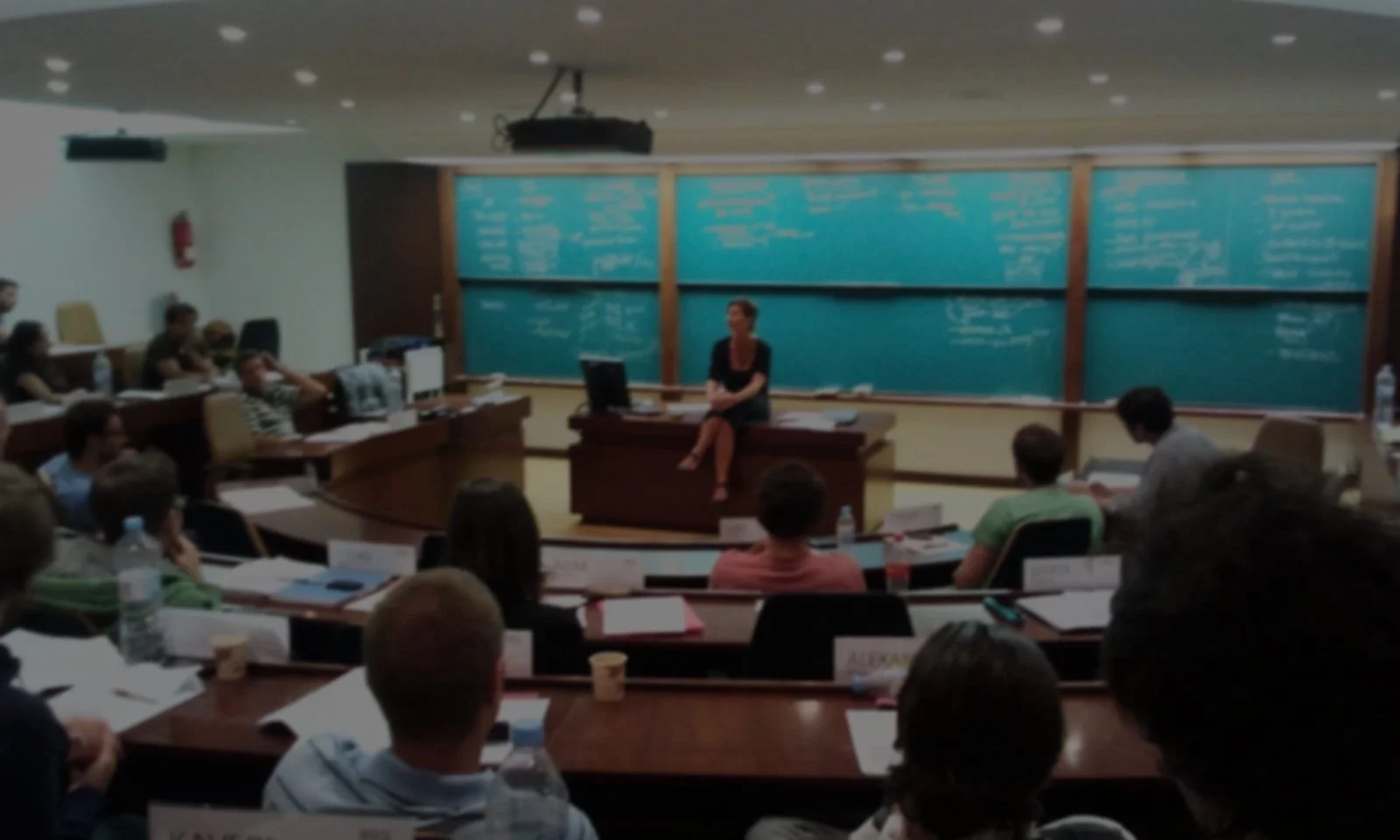 Een docent die voor een groot schoolbord met veel schriftuurlijke inhoud zit, terwijl een klas vol studenten op een gietvloer in een klaslokaal naar haar kijkt.