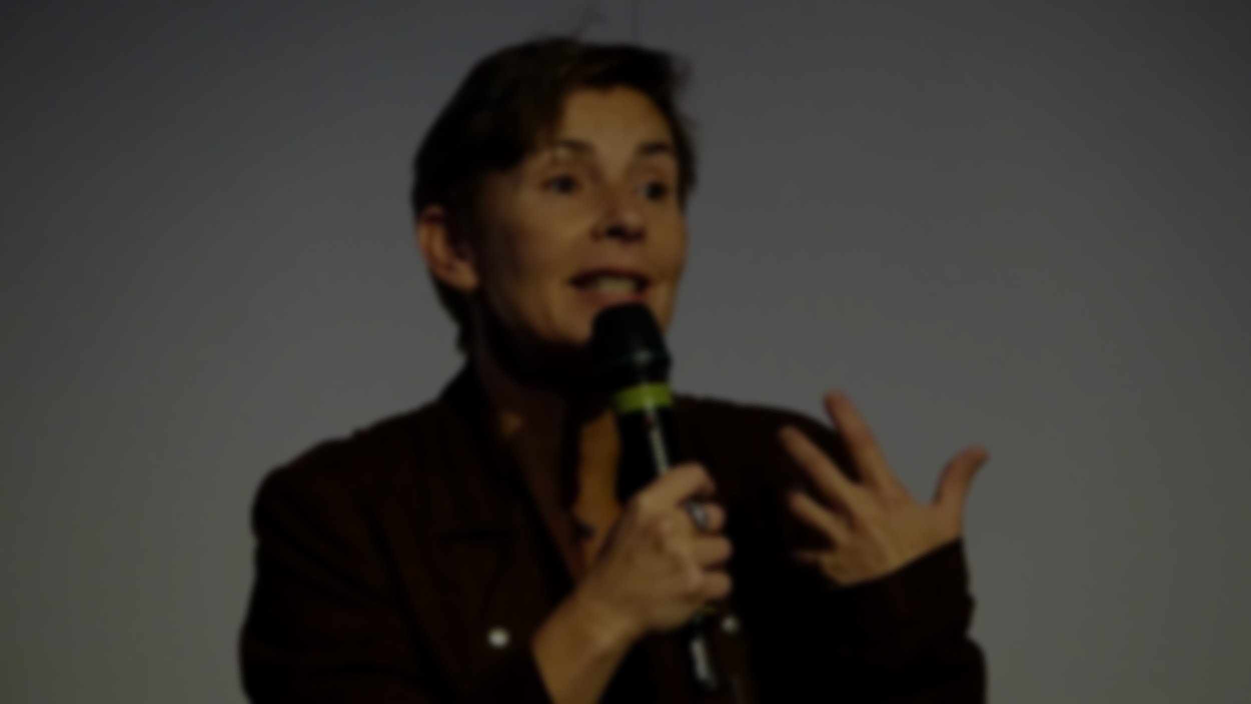 Inge Geerdens met microfoon tijdens presentatie, hand op borst, donkere achtergrond.
