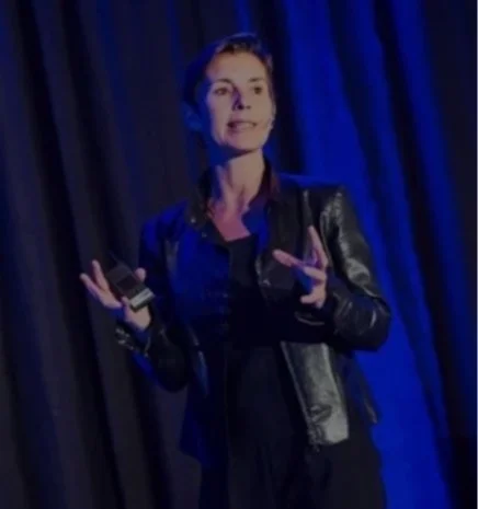 Inge Geerdens op podium die spreekt, met een microfoon en handen beweegt, blauwe achtergrond.