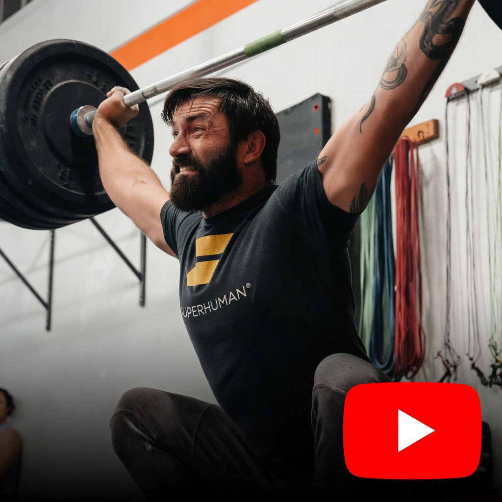 A man with a beard and tattoos lifting a barbell overhead during a workout at a gym.