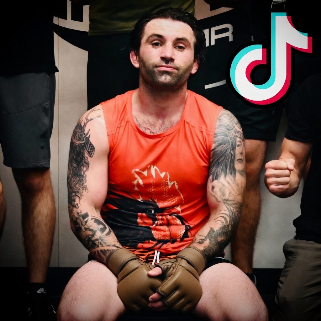 A man with tattoos on his arms and chest sitting down, wearing orange athletic gear and boxing gloves, surrounded by people, with a TikTok logo in the background.