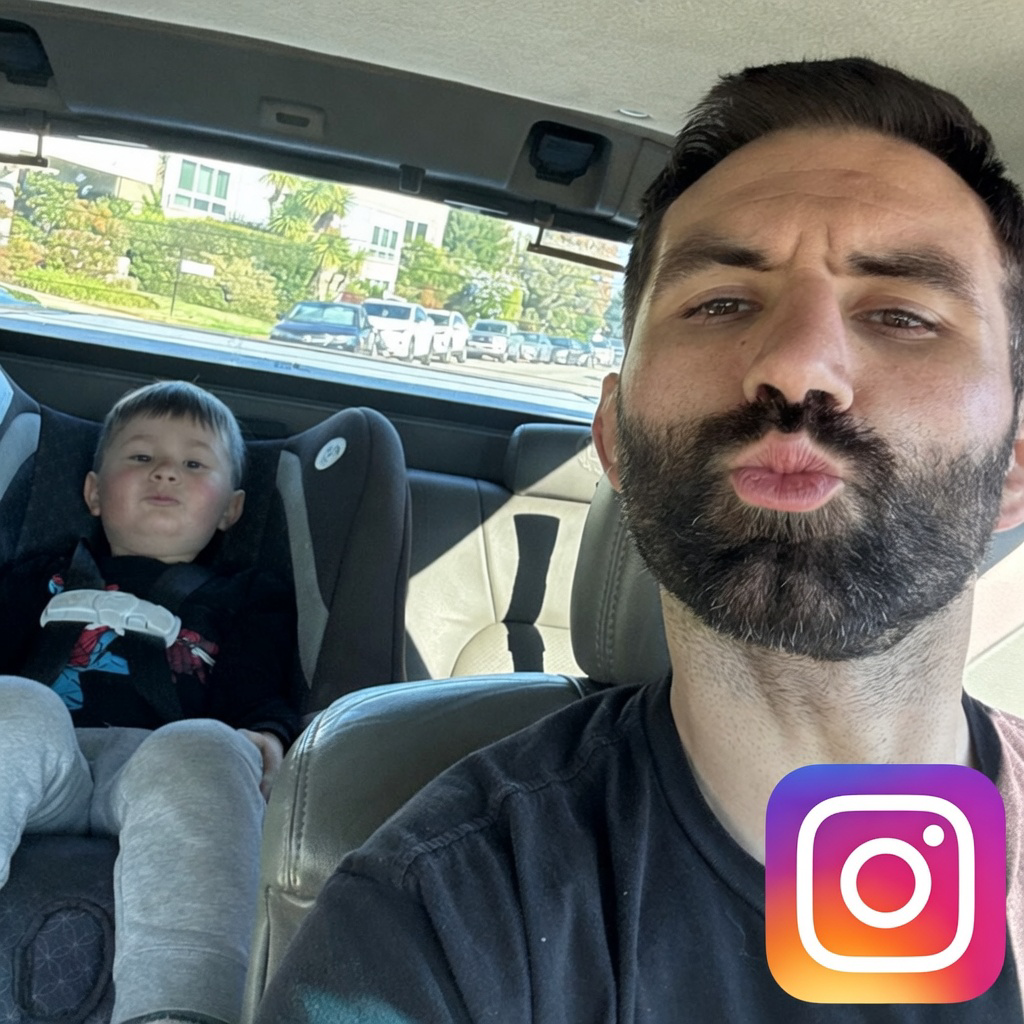A man with dark hair and a beard taking a selfie in the front passenger seat of a car, with a young boy sitting in a car seat behind him making a puckering face. The background shows a parking lot and residential buildings outside the car window.