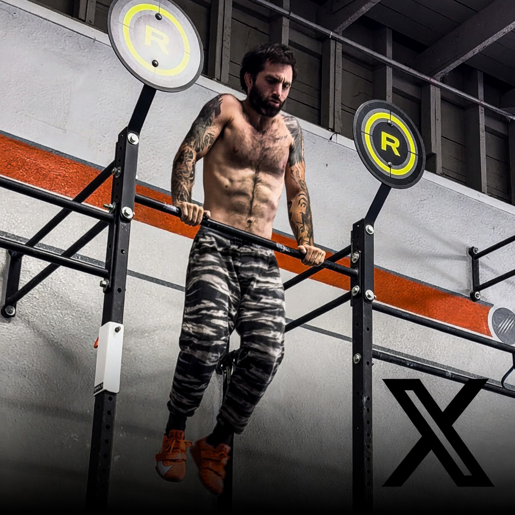 A man with tattoos on his arms and chest doing a dip exercise on a parallel bar in a gym. The gym has a gray wall with orange accents and two target signage with a yellow 'R' in a circle.