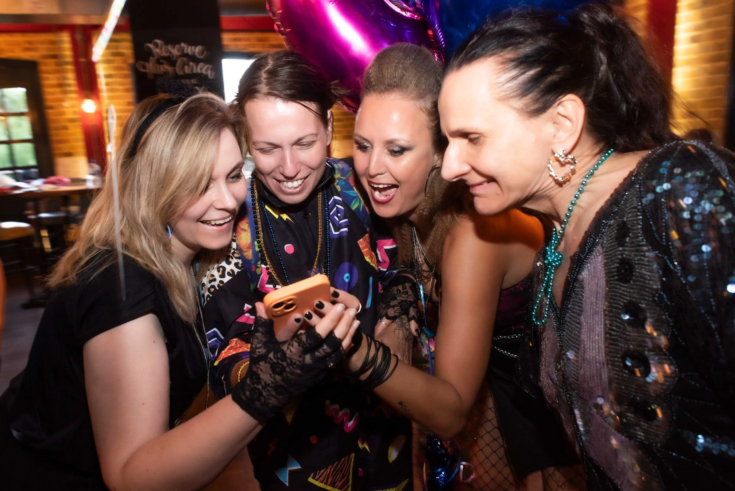 Group of women gathered around smiling and looking at a smartphone, celebrating at a party with balloons in the background.