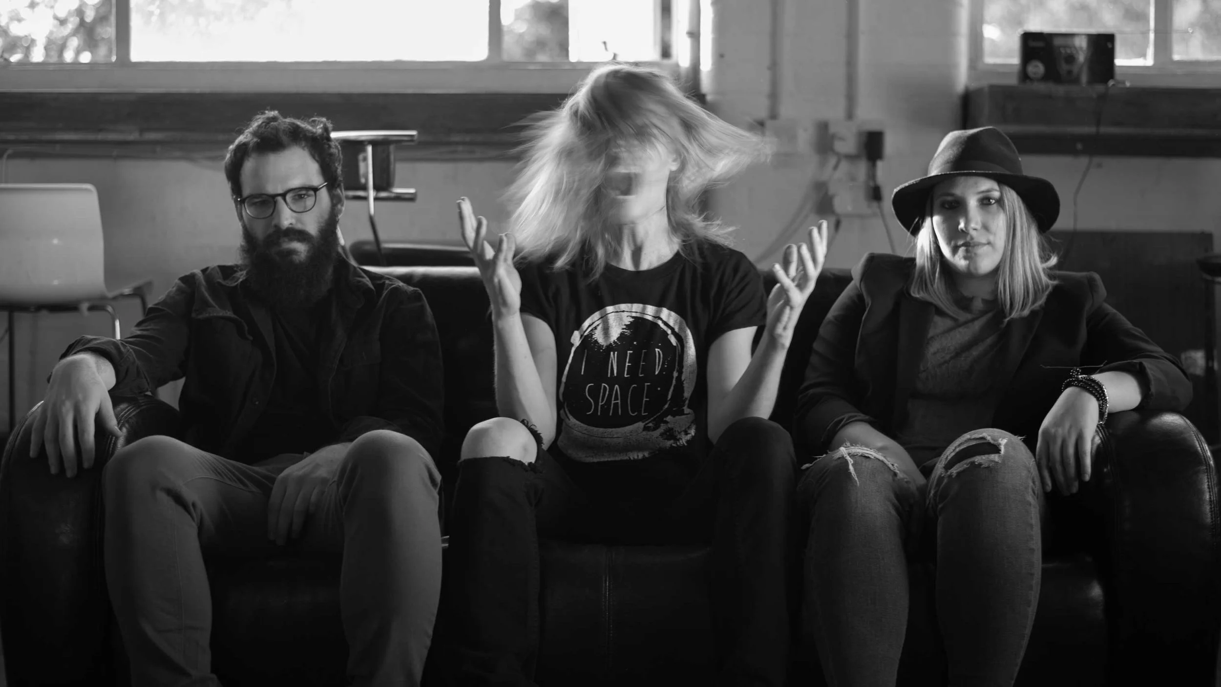 Neverlanded - Shot by Shotison. Black and white photo of three young adults sitting on a leather couch, with a man in the middle flipping hair, a man with glasses and a beard on the left, and a woman wearing a hat on the right.