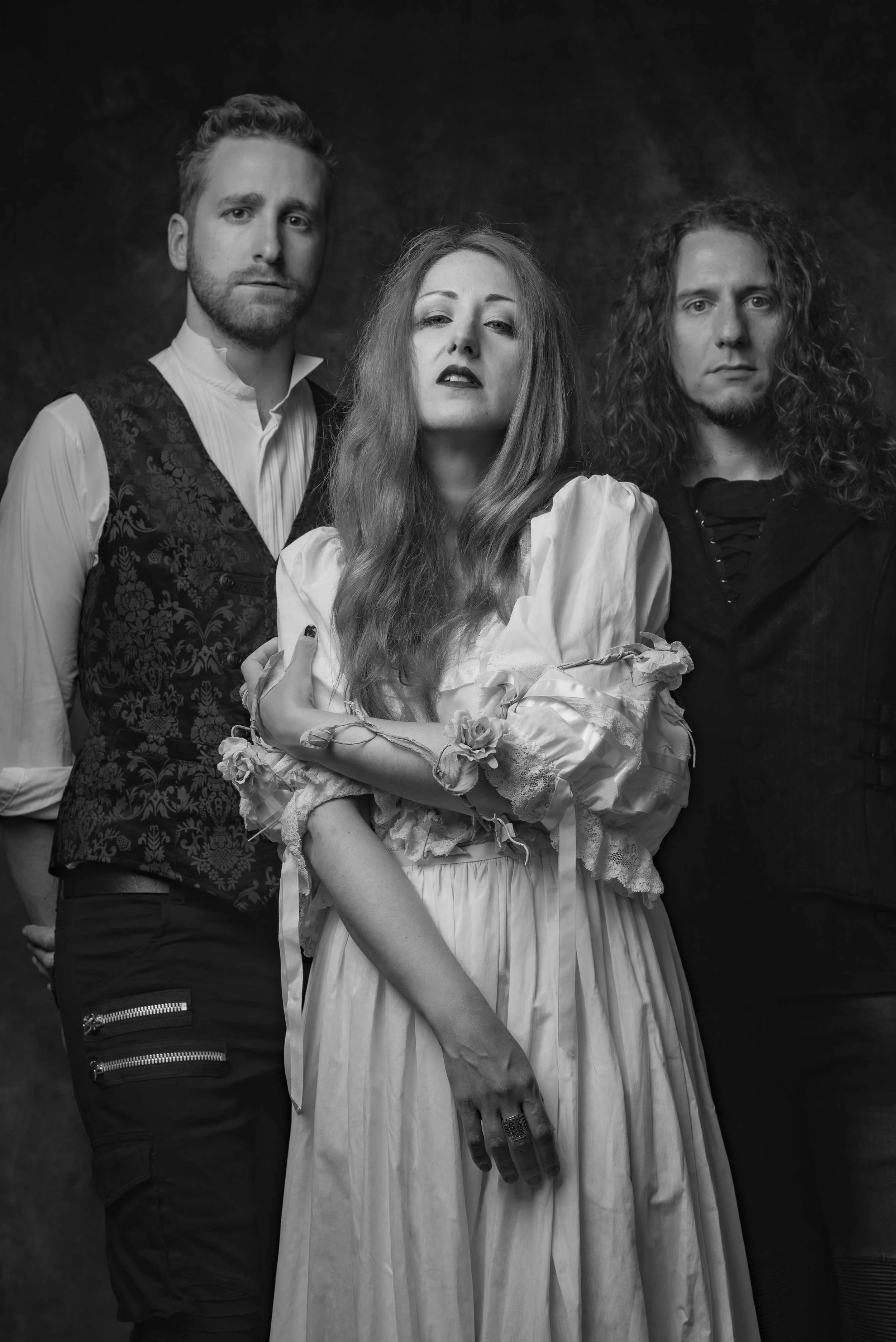Alice is Sleeping - Shot by Shotison. Three people: a man with short hair and a beard on the left, a woman with long wavy hair and makeup in the center, and a man with curly long hair on the right, standing against a dark background.