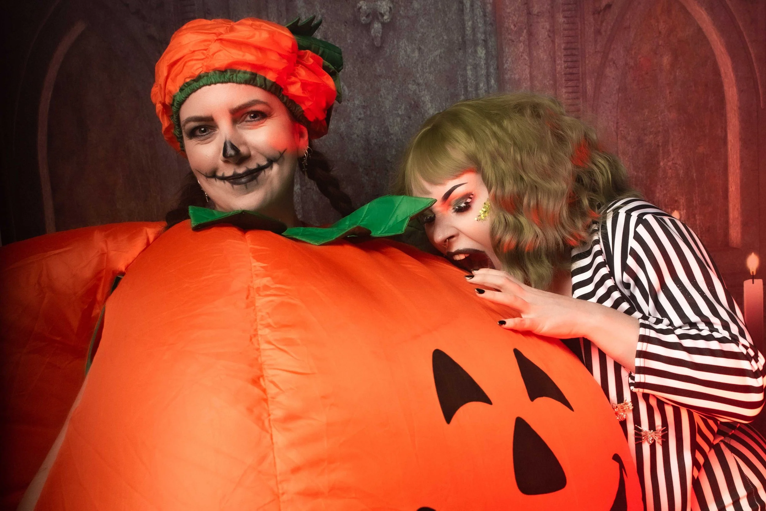 Two women in Halloween costumes, one dressed as a pumpkin with face paint, the other with Beetlejuice green hair and makeup, leaning on a large inflatable pumpkin with a jack-o'-lantern face.