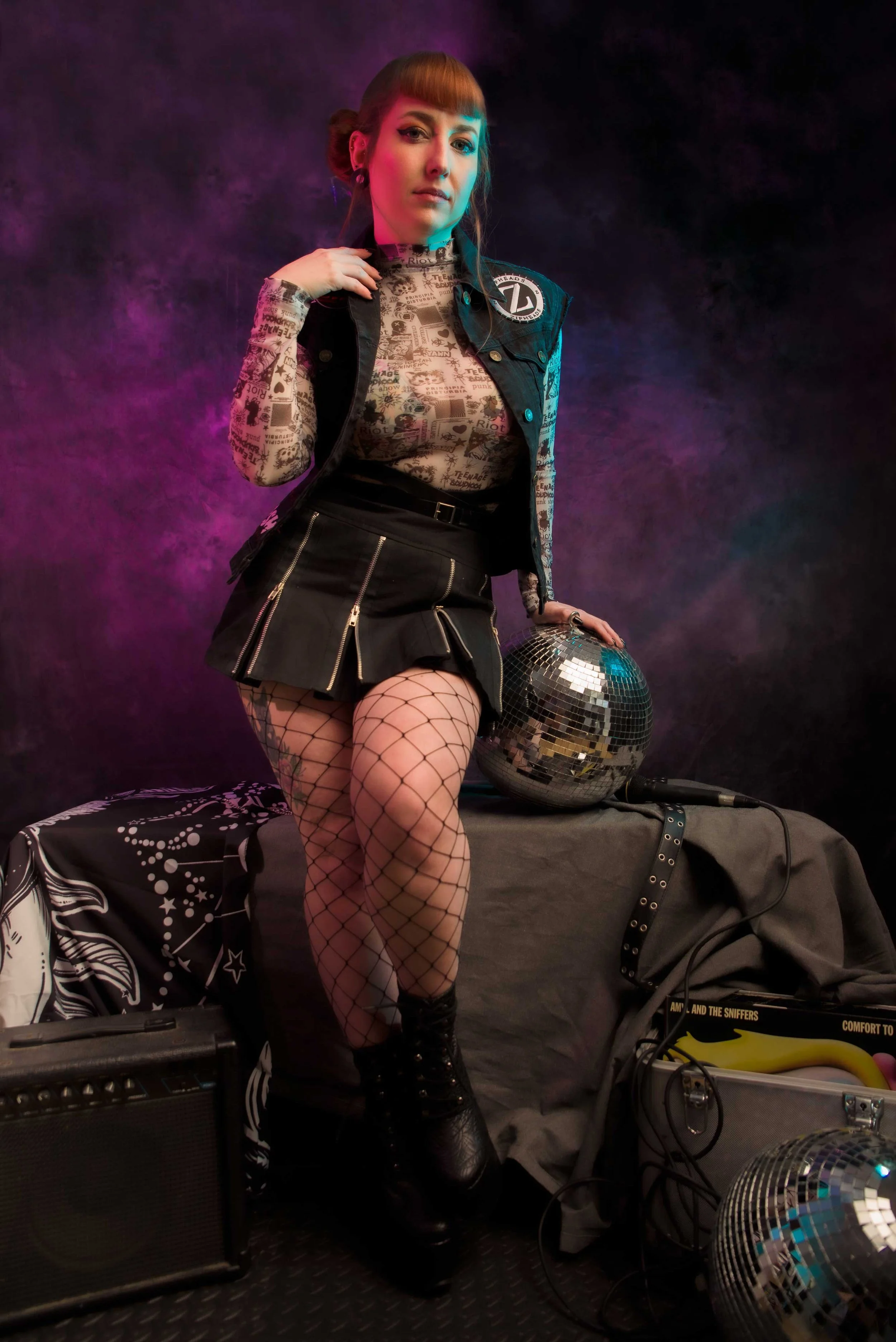 A woman with red hair, wearing fishnet stockings, black combat boots, a black skirt with zippers, and a sheer shirt with text and graphic designs, poses in a dark, smoky setting. She is leaning on a table covered with a black cloth, surrounded by musical equipment, including an amplifier and disco balls. She has a confident expression and is slightly squinting, with her hand near her neck.