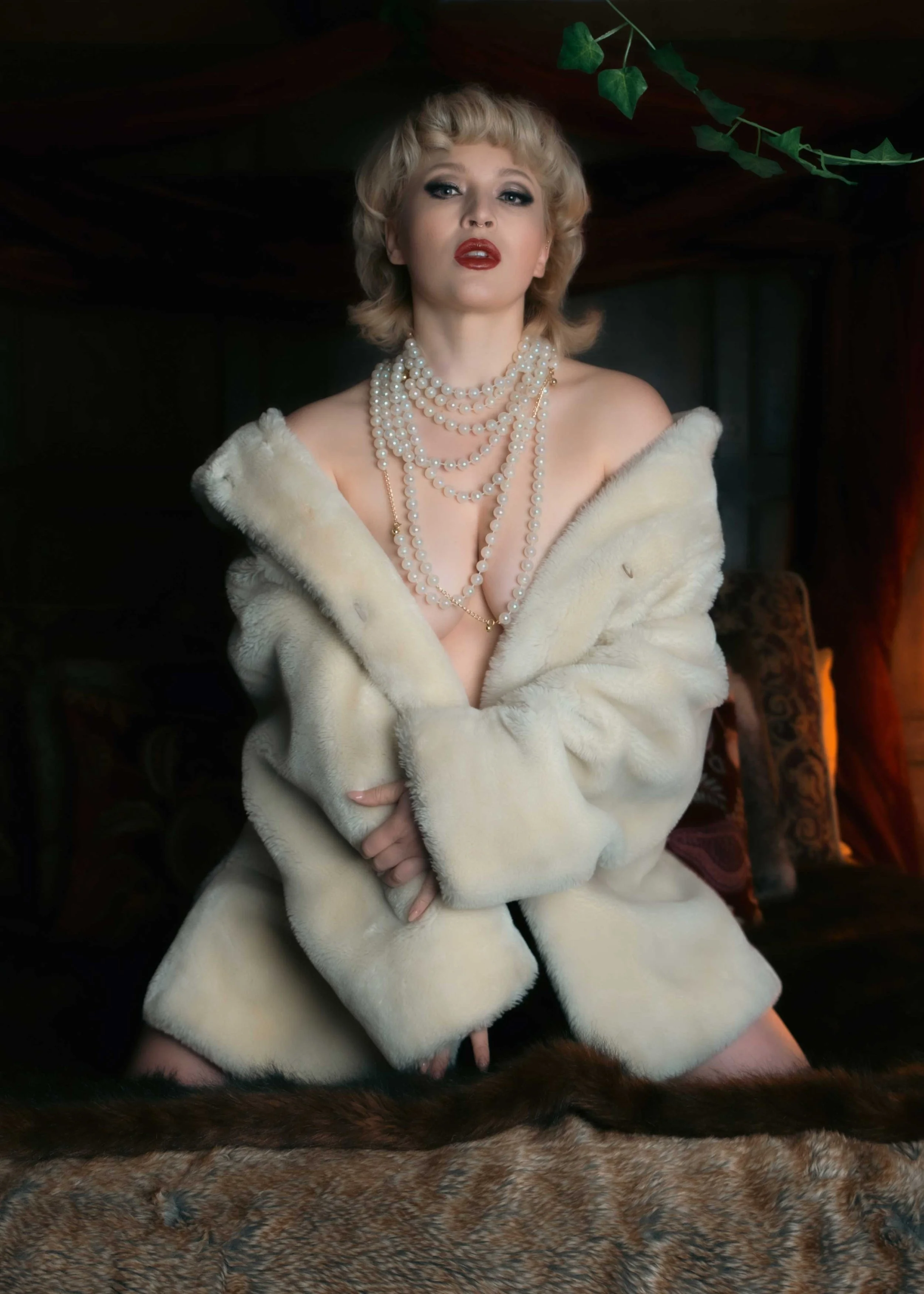 A woman with blonde hair and makeup wearing a white fur coat and multiple strands of pearl necklaces, sitting on a patterned rug with dark background and some green foliage.