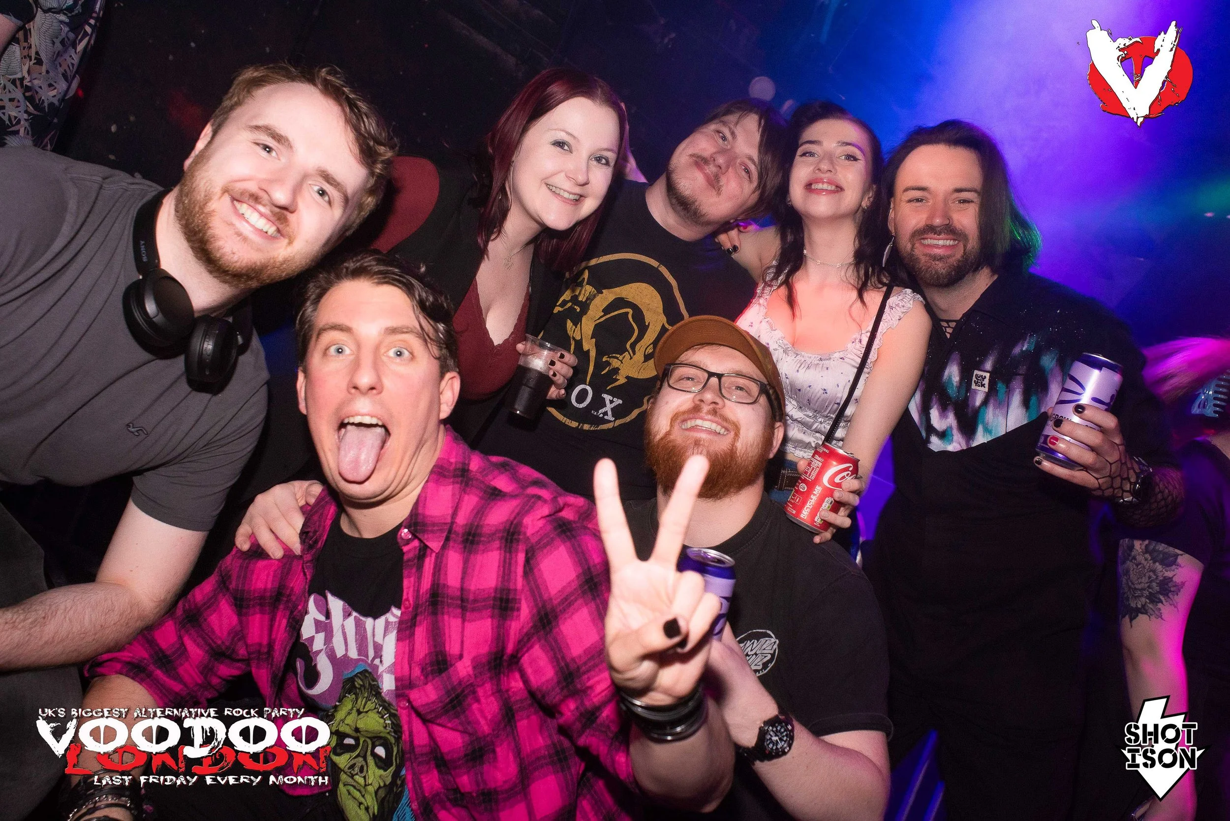 Group of six people smiling and posing at a party, some holding drinks and making peace signs, in a dimly lit venue with colorful lights and a logo in the corner.