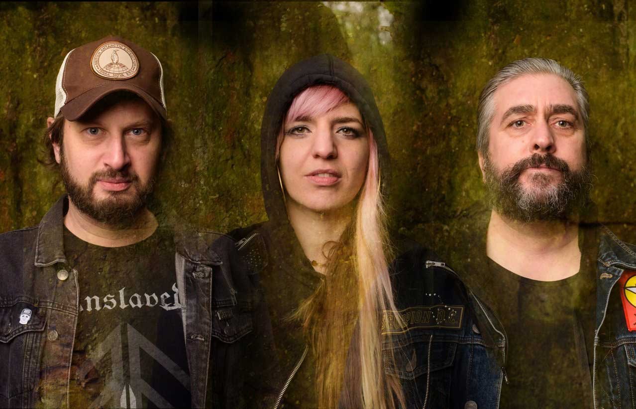 Possessor - Shot by Shotison. Three people standing in front of a forest background, dressed in dark clothing with patches and graphic T-shirts, looking directly at the camera.