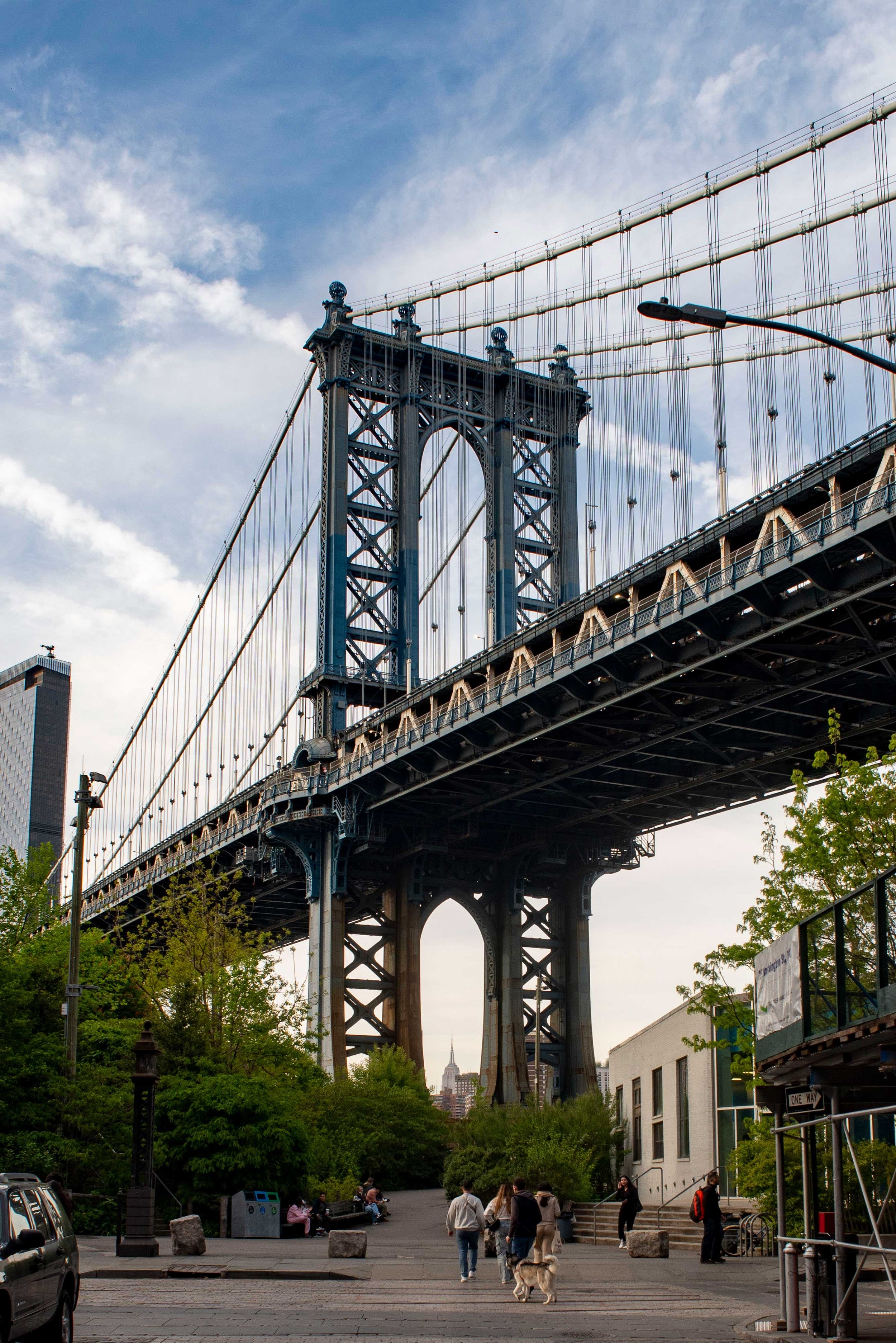 View of the Brooklyn Bridge over the East River in New York City with pedestrians walking underneath and trees lining the pathway. The Empire State Building is visible in the background.