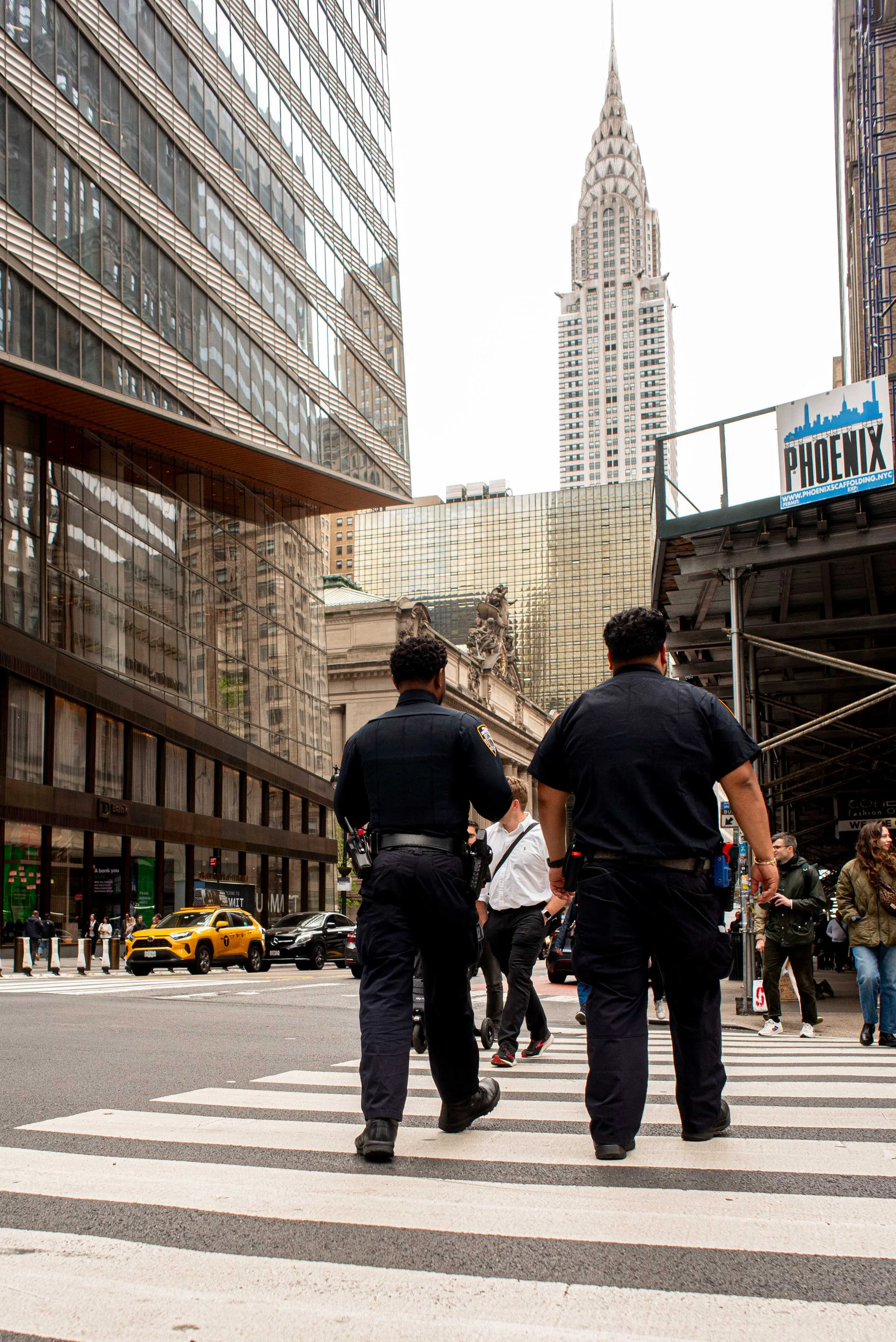 Two police officers walking across a busy city street at a crosswalk with tall buildings and the Chrysler building in the background, with numerous pedestrians and taxis on the street.