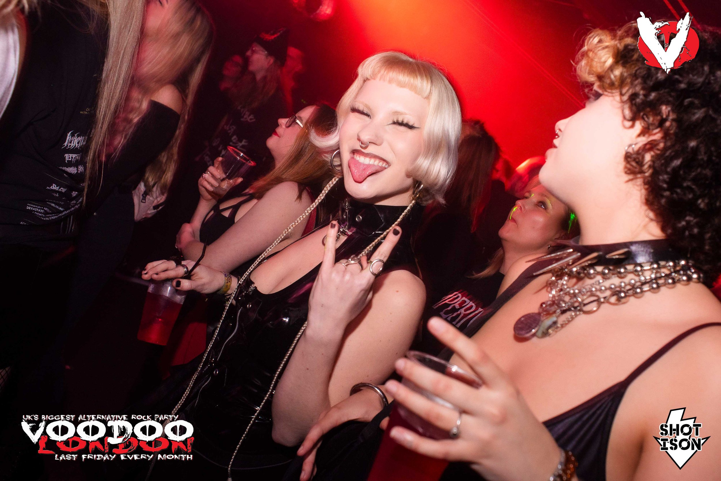 Young woman with platinum blonde hair, piercings, and makeup smiling and sticking out her tongue at a party with red lighting, surrounded by other women.