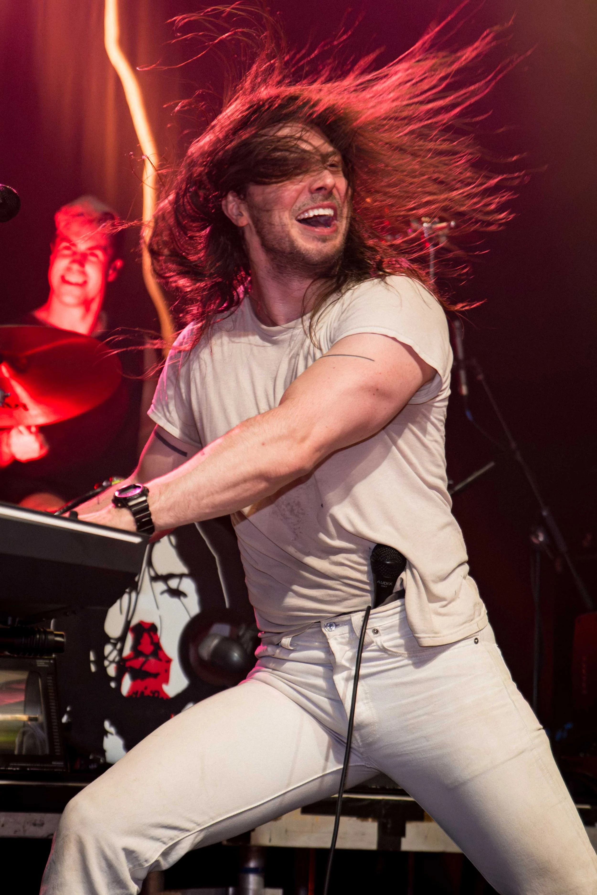 Andrew WK - Shot by Shotison. A man with long dark hair, wearing a beige t-shirt and white pants, intensely playing keyboard on stage with his hair flying in the air, and smiling. In the background, a drummer is visible, also smiling.