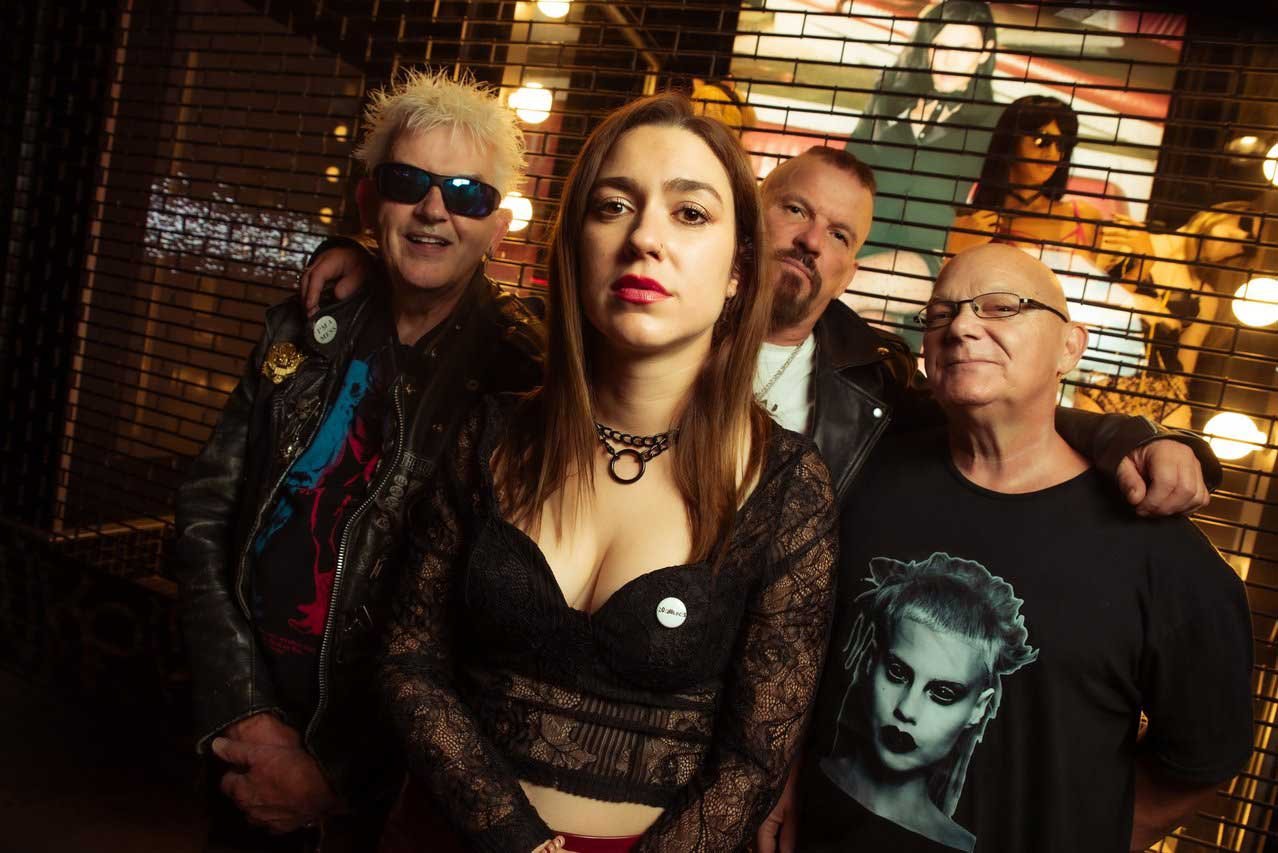 No Feelings - Shot by Shotison. Four people posing in front of a decorative wall with lighting and images of musicians, with a bar or club-like setting.