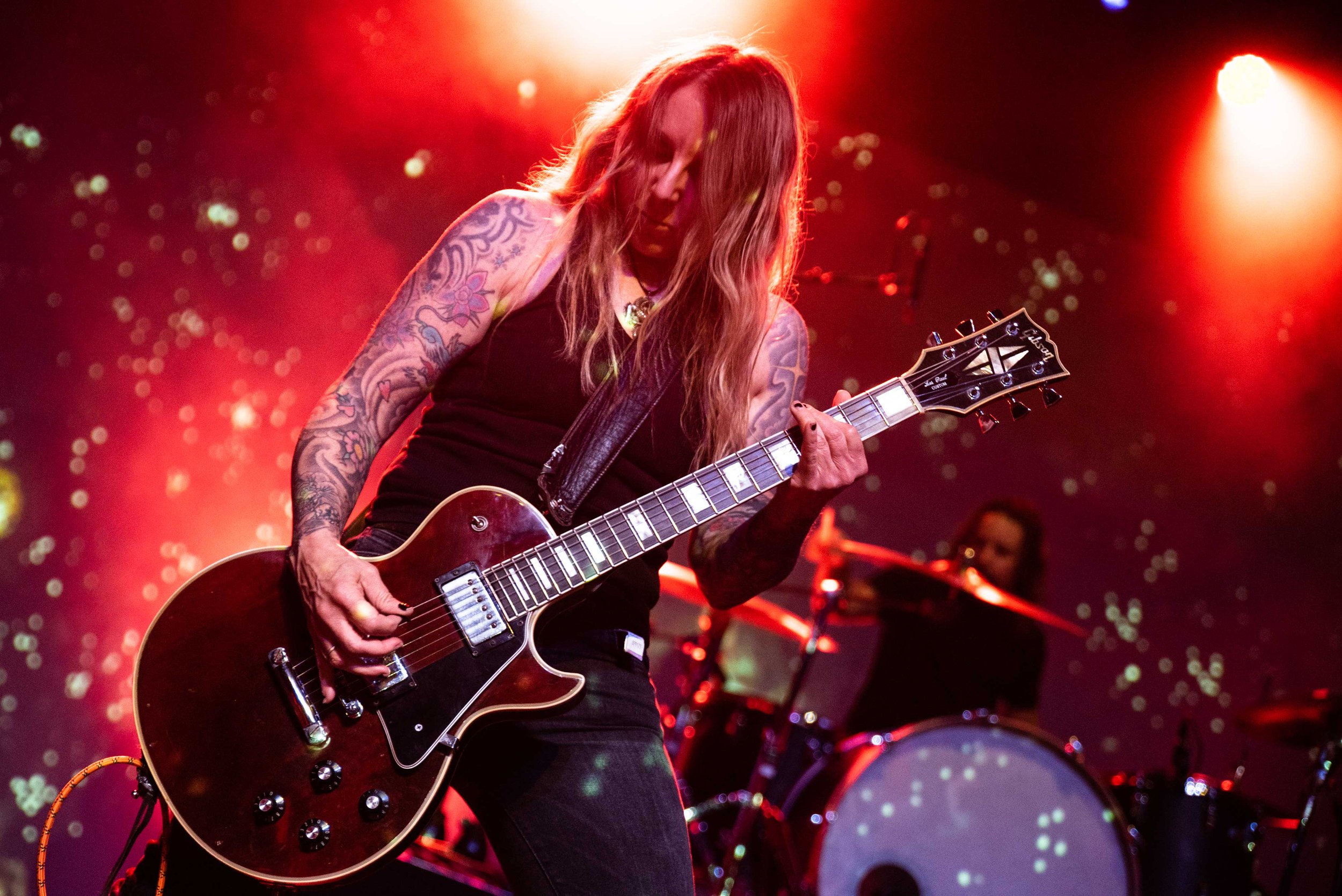 Acid King - Shot by Shotison. A female musician with long hair and tattoos playing an electric guitar on stage under red and orange lighting.