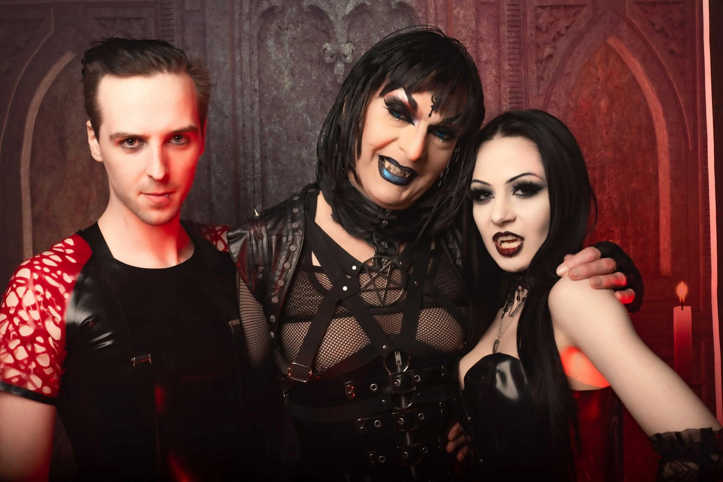 Three individuals dressed in gothic or punk fashion, standing close together in a dimly lit room with gothic decor. The person on the left has short hair, pale skin, and is wearing a mesh top with red accents. The person in the middle has black hair, heavy makeup with blue lipstick and eye makeup, and is wearing a black leather jacket and mesh top. The person on the right has long black hair, dark makeup, and is dressed in black clothing with gothic accessories. They are posing together with confident expressions.