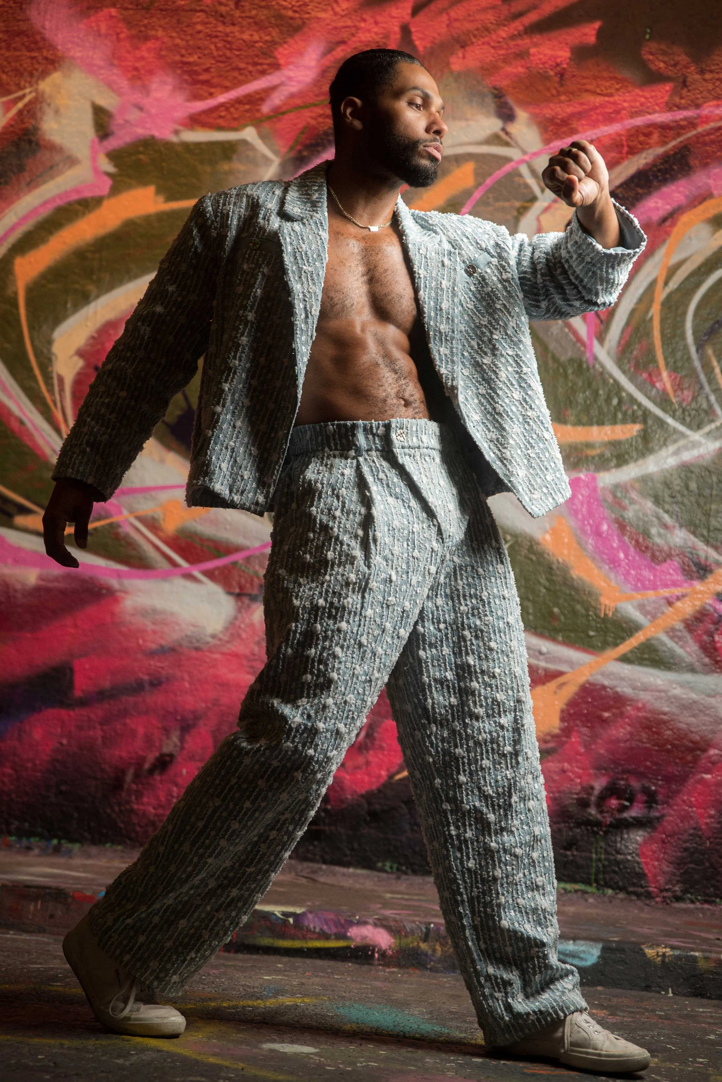 Man wearing a textured suit with no shirt underneath, standing against a colorful graffiti background, in a dynamic pose with one arm bent and fist near his face, and wide-legged stance.