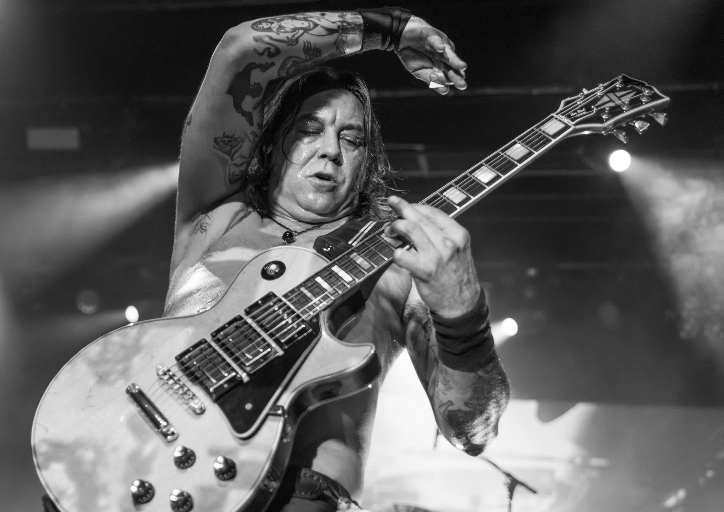 A male guitarist with long hair and tattoos plays an electric guitar during a live performance, with a dark and smoky stage background.