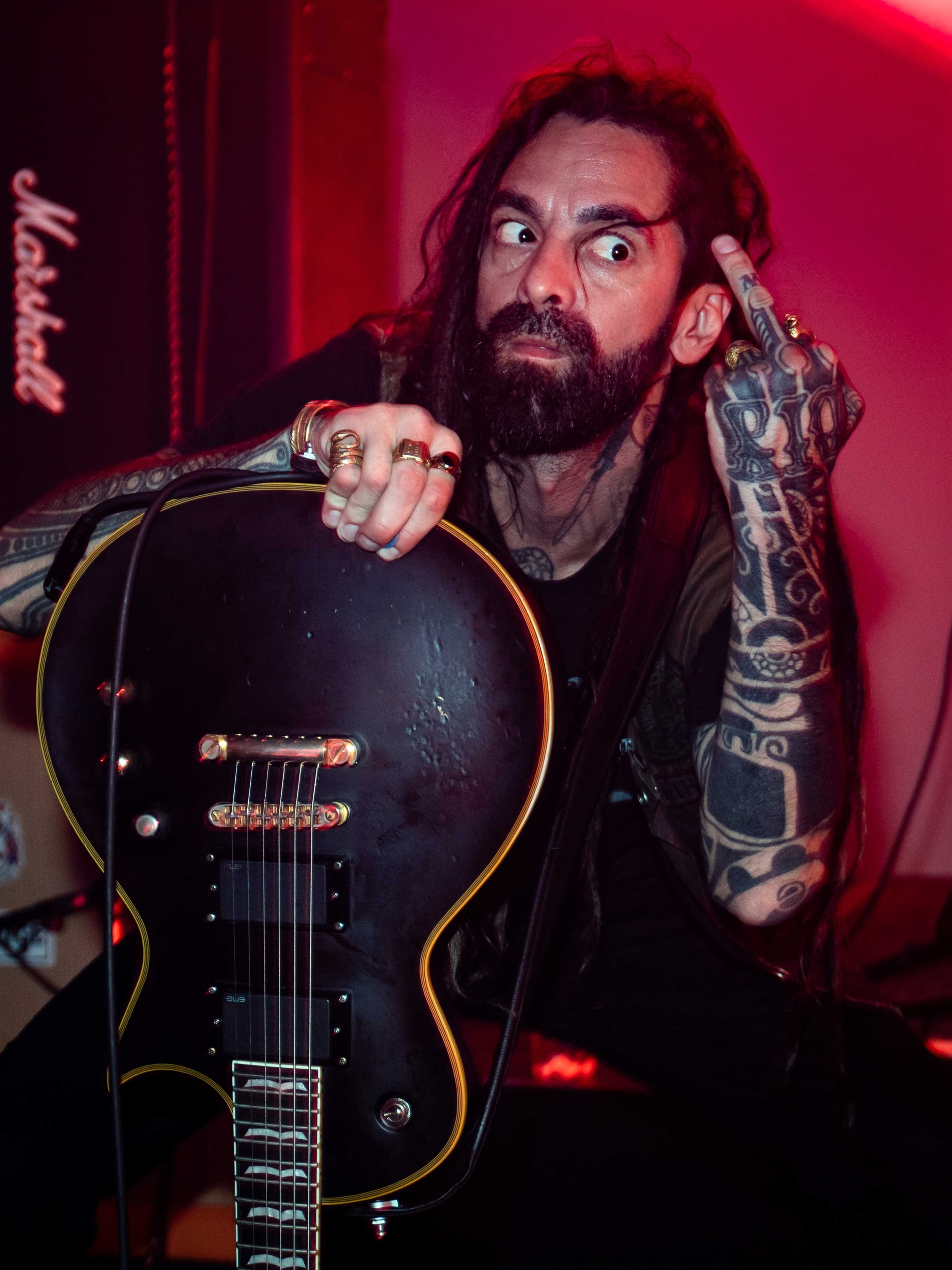 Dopethrone - Shot by Shotison. A man with long dark hair and tattoos, wearing rings, holding an electric guitar, sitting in a dimly lit room with red lighting, making a gesture with his fingers, with a surprised or intense facial expression.