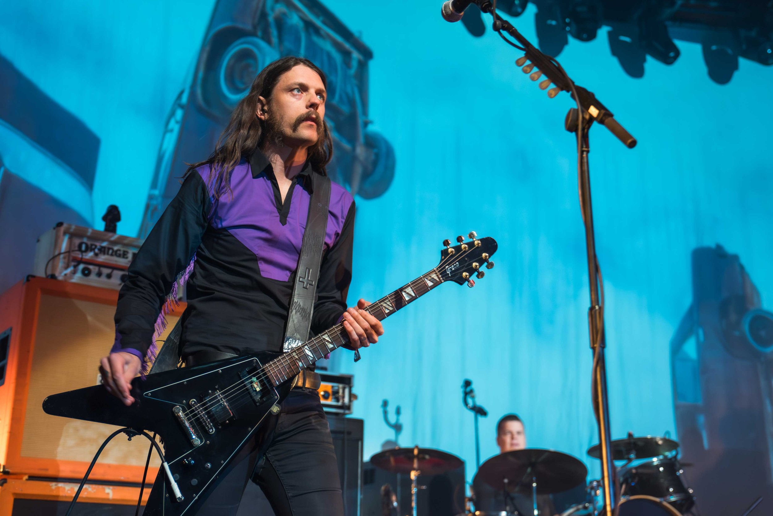Monolord - Shot by Shotison. A musician with long dark hair and a moustache playing a black electric guitar on stage, wearing a purple and black shirt, with a microphone stand nearby and other band members in the background.