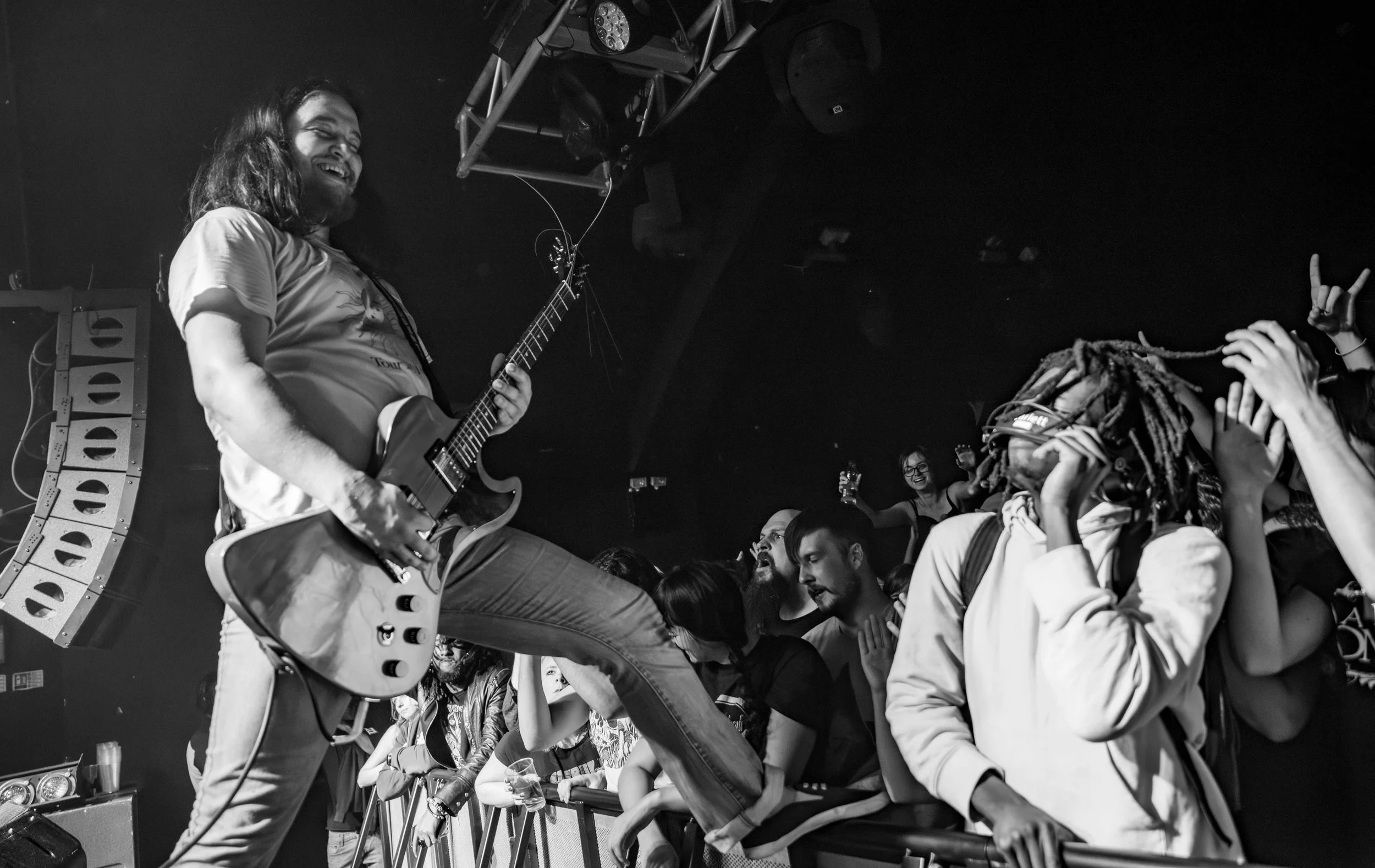 Stoned Jesus - Shot by Shotison. Guitarist on stage playing an electric guitar at a concert, with a lively crowd reaching towards the stage and cheering. The scene is black and white, capturing the energy and excitement of the live music performance.