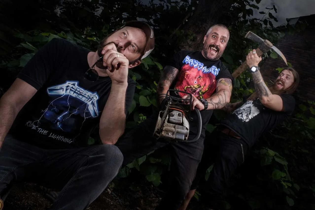 Possessor - Shot by Shotison. Three men standing outdoors, one holding a chainsaw, one holding a sledgehammer, and the third with a hand on his mouth, all smiling, in front of green foliage.