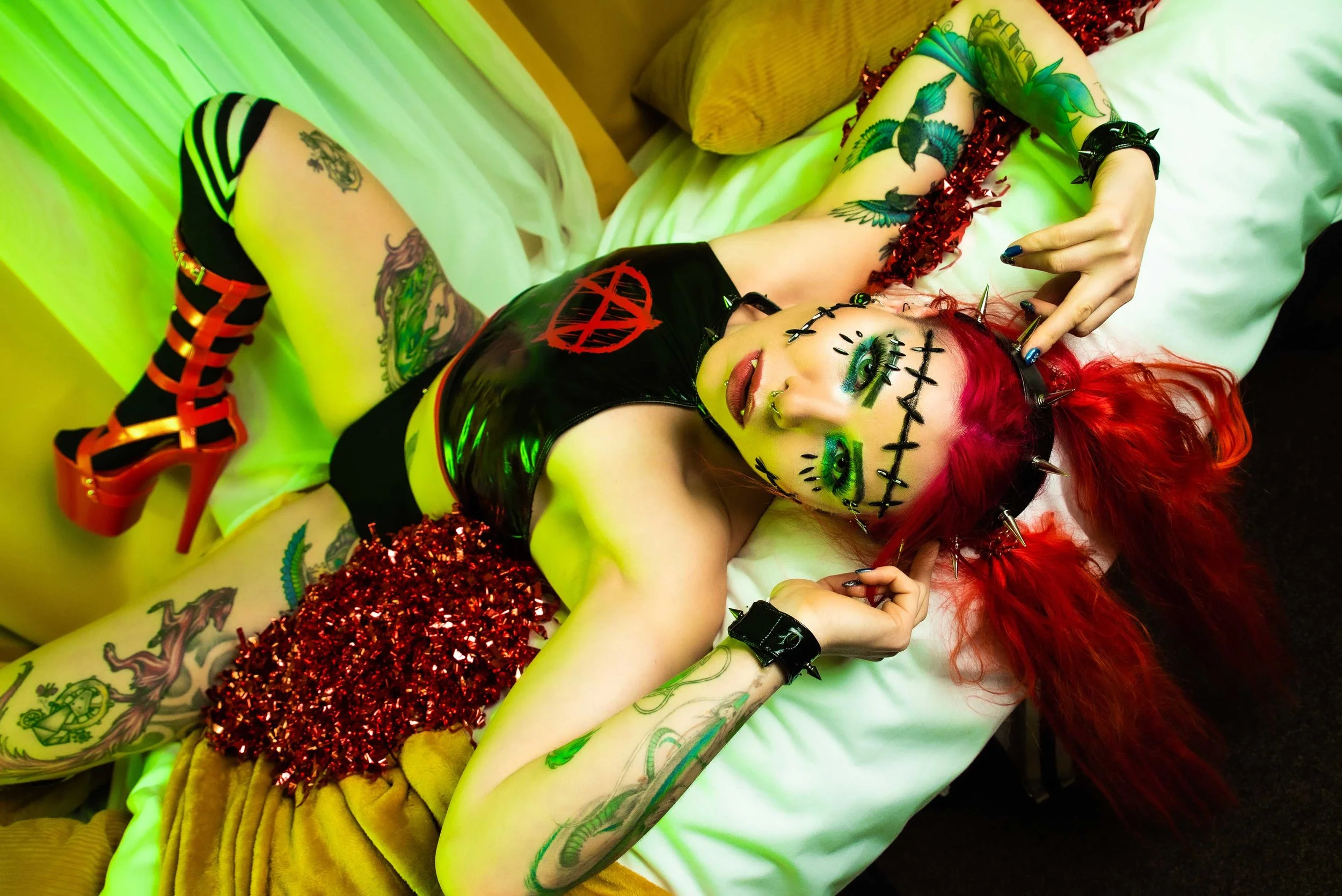 A person with bright red hair and colorful makeup lying on a bed. They have tattoos on their arms and legs and are wearing a black latex top, red and black striped boots, and a black cap with spikes. They are surrounded by red glitter and green lighting.