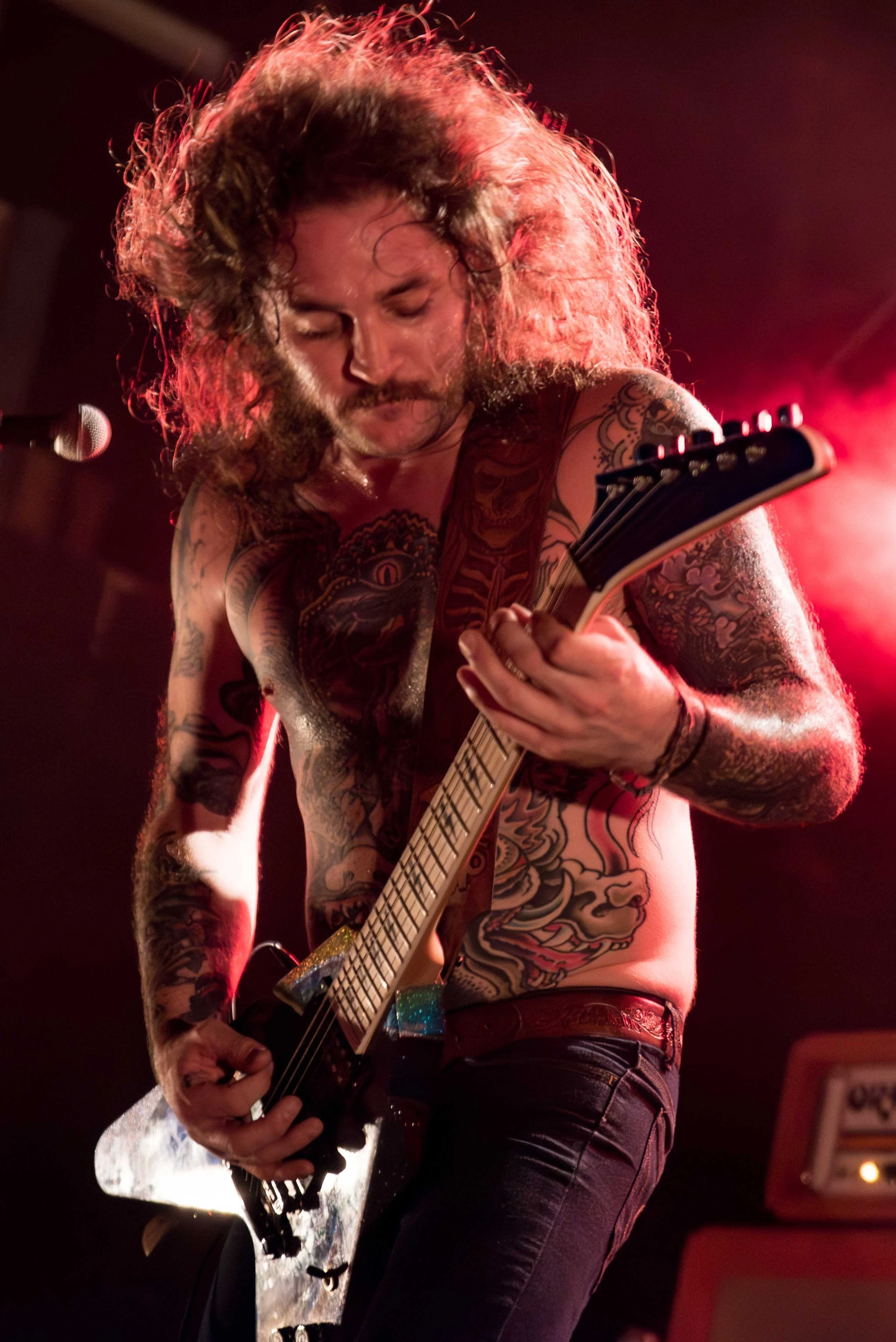 Mothership - Shot by Shotison. A shirtless man with long hair and tattoos on his arms and torso playing an electric guitar on stage under red lighting.